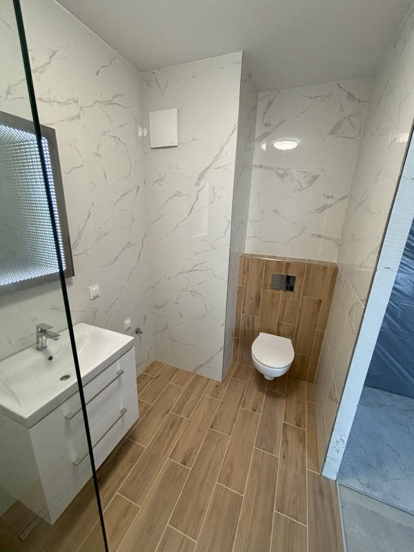 Wykończona łazienka z białymi płytkami imitującymi marmur i drewnopodobną podłogą. Umywalka z szafką i toaleta w zabudowie. Widok z kabiny prysznicowej.