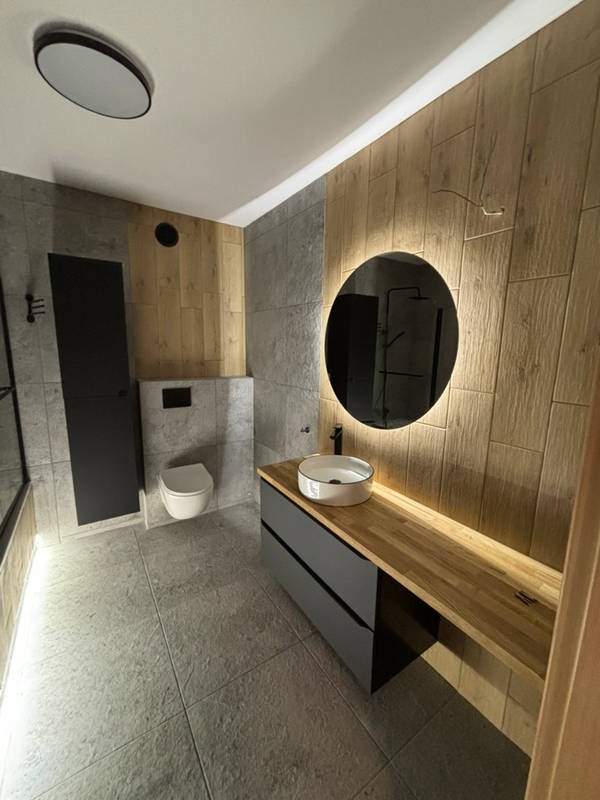 Nowoczesna łazienka z drewnianymi i szarymi płytkami, okrągłym lustrem z podświetleniem LED, umywalką nablatową i szafką z szufladami. Minimalistyczny design.