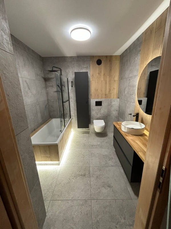 Nowoczesna łazienka z szarymi płytkami, drewnianymi akcentami i podświetlaną wanną. Okrągłe lustro z podświetleniem nad umywalką.