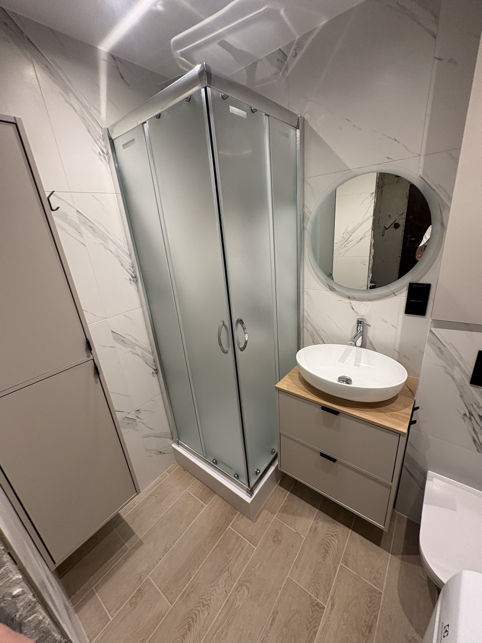 Nowoczesna łazienka z kabiną prysznicową, umywalką na szafce z drewnianym blatem i okrągłym lustrem. Marmurowe płytki na ścianach i drewnopodobna podłoga.