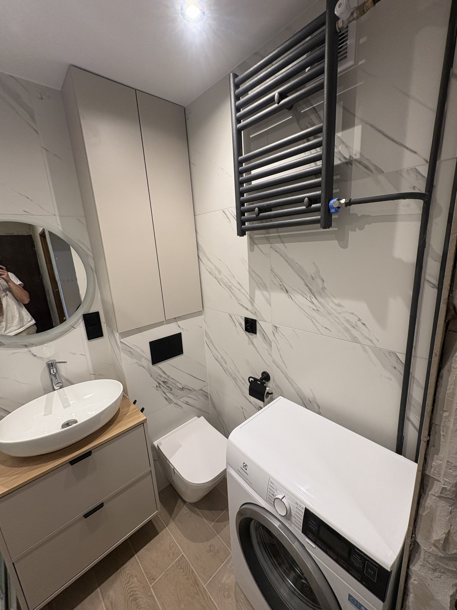 Nowoczesna łazienka z marmurowymi płytkami, umywalką na szafce, toaletą, grzejnikiem drabinkowym i pralką Electrolux. Minimalistyczny design, jasne kolory.