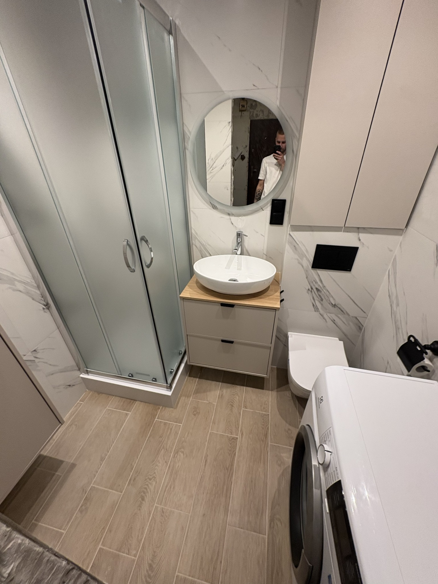 Nowoczesna łazienka z kabiną prysznicową, okrągłym lustrem i umywalką nablatową. Beżowe płytki podłogowe i marmurowe na ścianach. Widoczna pralka i toaleta.