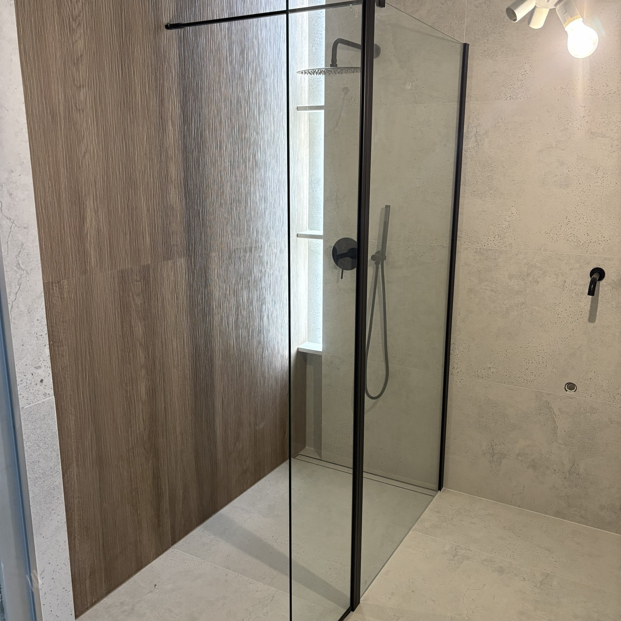 Nowoczesna kabina prysznicowa walk-in z czarnymi profilami, drewnopodobne płytki na ścianie i szara armatura w wykończonej łazience. Minimalistyczny design.