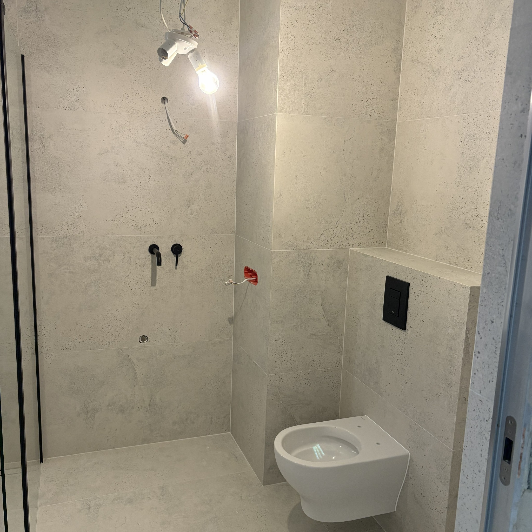 Wykończona łazienka w szarych płytkach imitujących beton. Widoczna toaleta, czarne baterie i fragment kabiny prysznicowej. Oświetlenie tymczasowe.