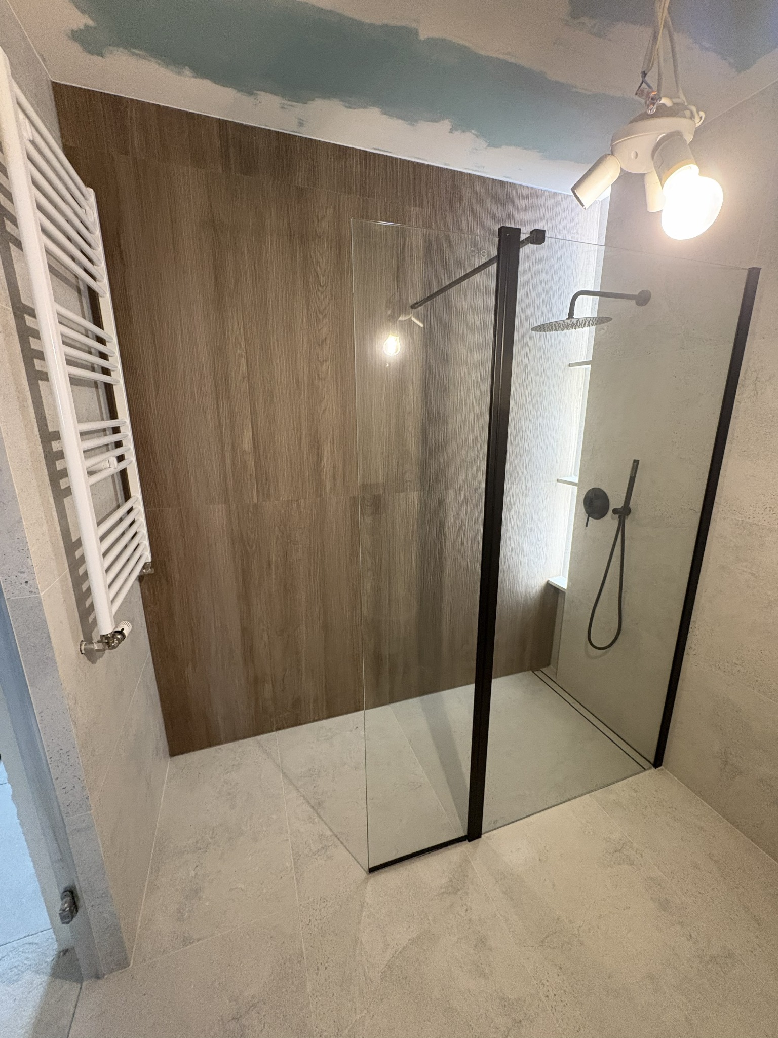 Nowoczesna kabina prysznicowa z czarnymi profilami, grzejnik drabinkowy i drewnopodobne panele ścienne w wykończonej łazience z sufitowym malunkiem chmur.