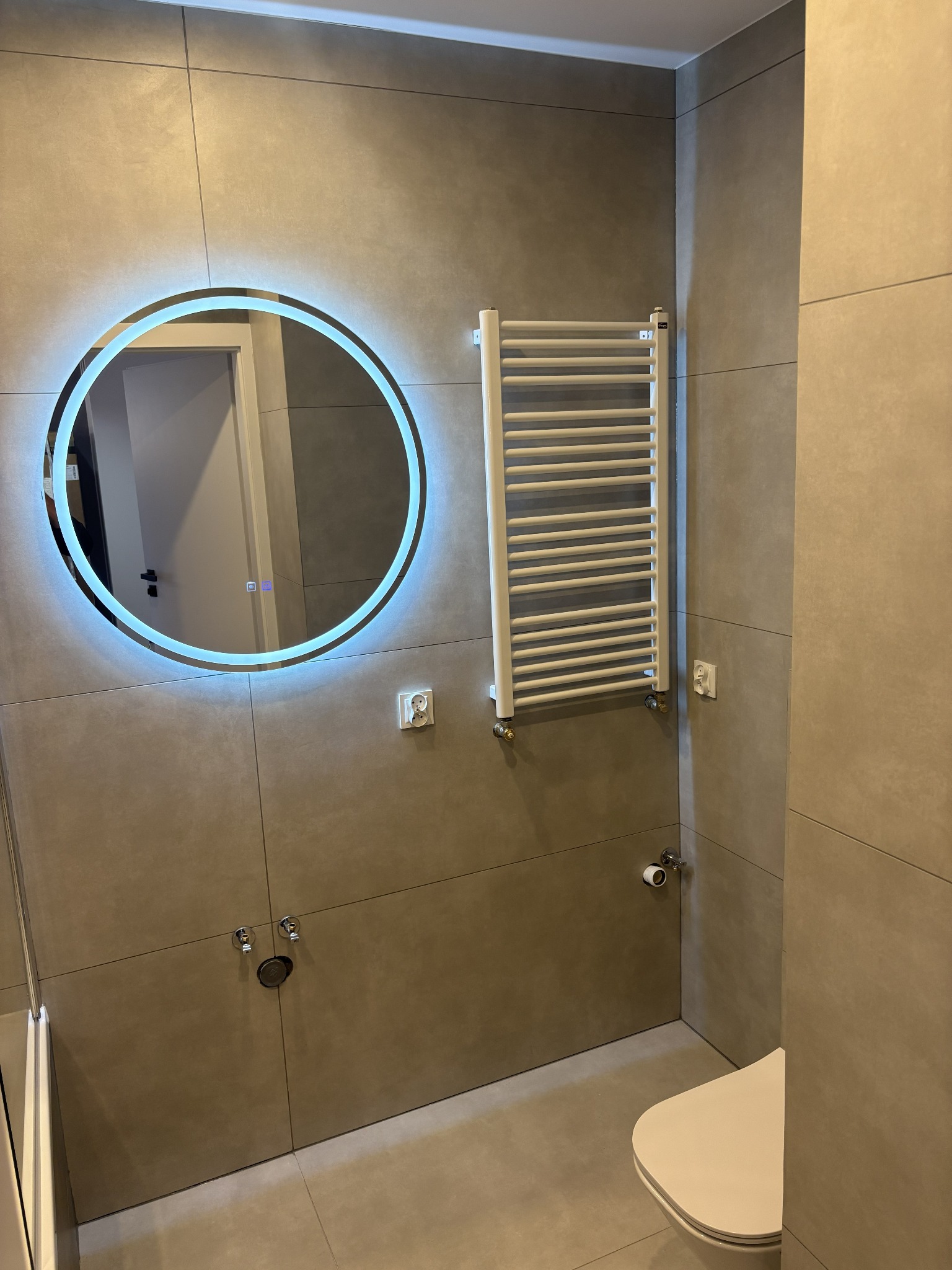 Wykończona łazienka z beżowymi płytkami, okrągłym lustrem z podświetleniem LED, białym grzejnikiem drabinkowym i toaletą. Minimalistyczny design.