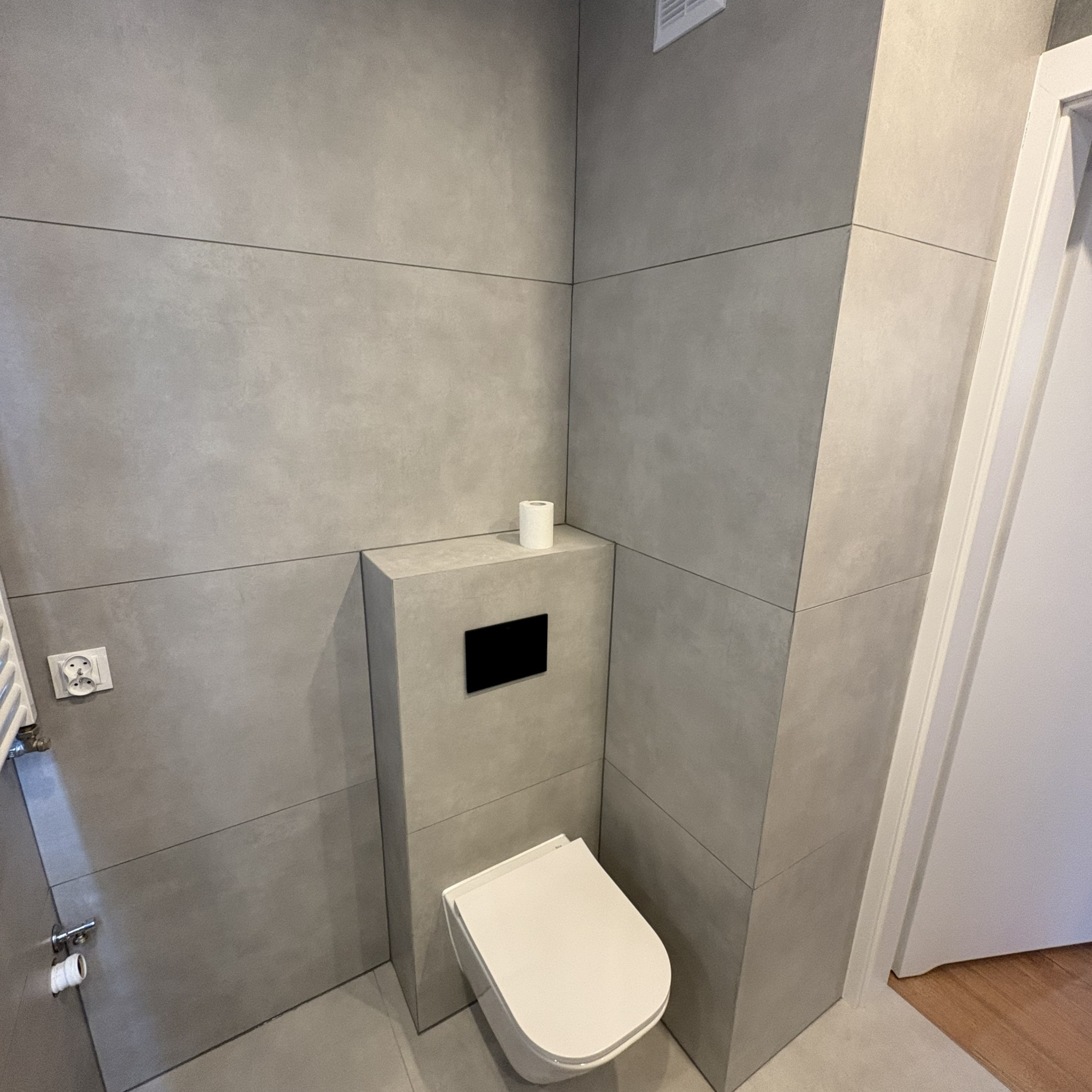 Nowoczesna łazienka z szarymi płytkami wielkoformatowymi, podwieszana toaleta z czarnym przyciskiem, minimalistyczny design, rolka papieru toaletowego.