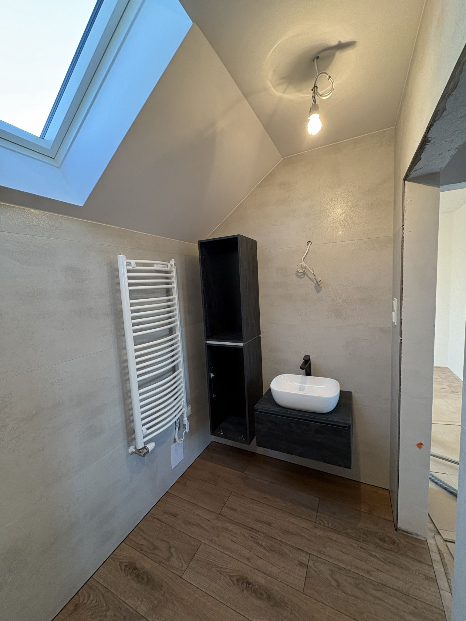 Wykończona łazienka na poddaszu z oknem dachowym. Umywalka na szafce, grzejnik drabinkowy i drewnopodobne płytki podłogowe. Widoczne niedokończone obramowanie drzwi.