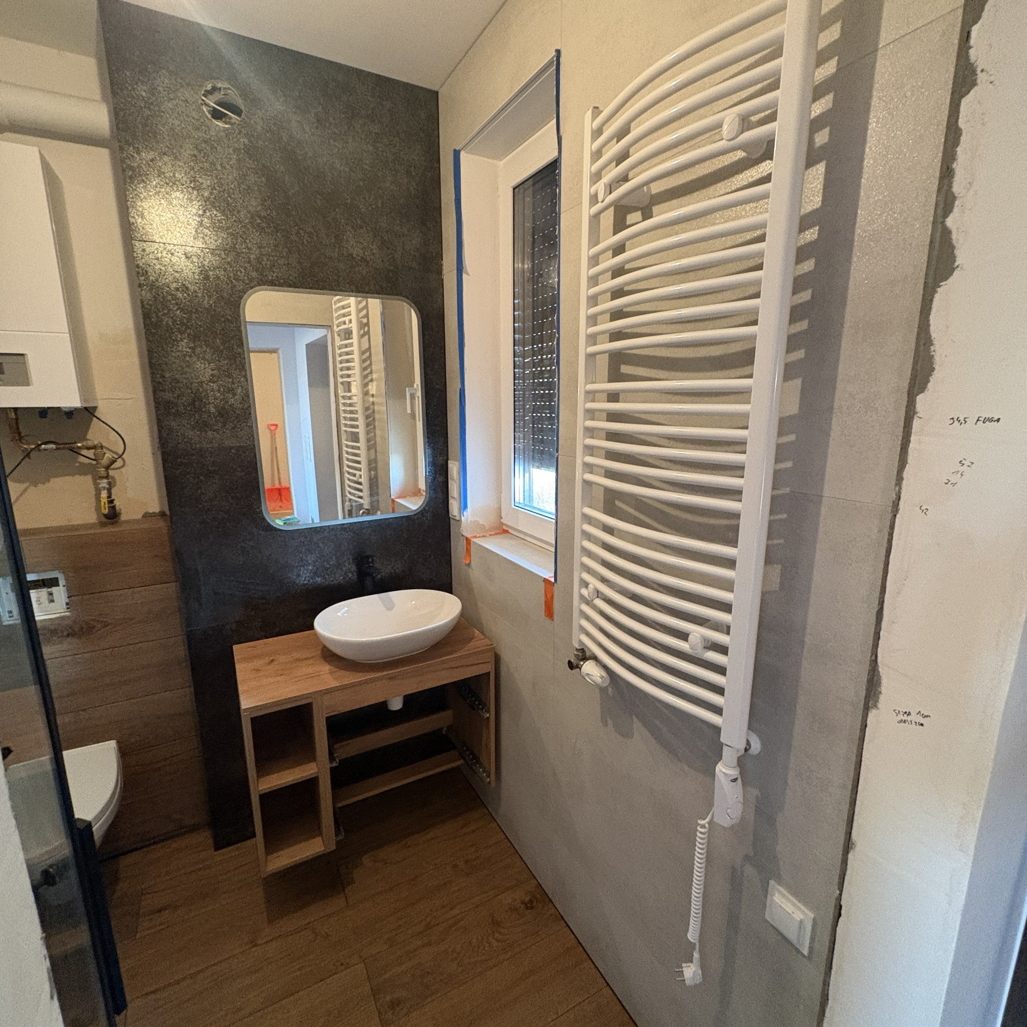 Łazienka w trakcie remontu: zamontowany grzejnik, umywalka na drewnianym blacie, lustro na tle ciemnej ściany. Widoczne ślady po pracach wykończeniowych.