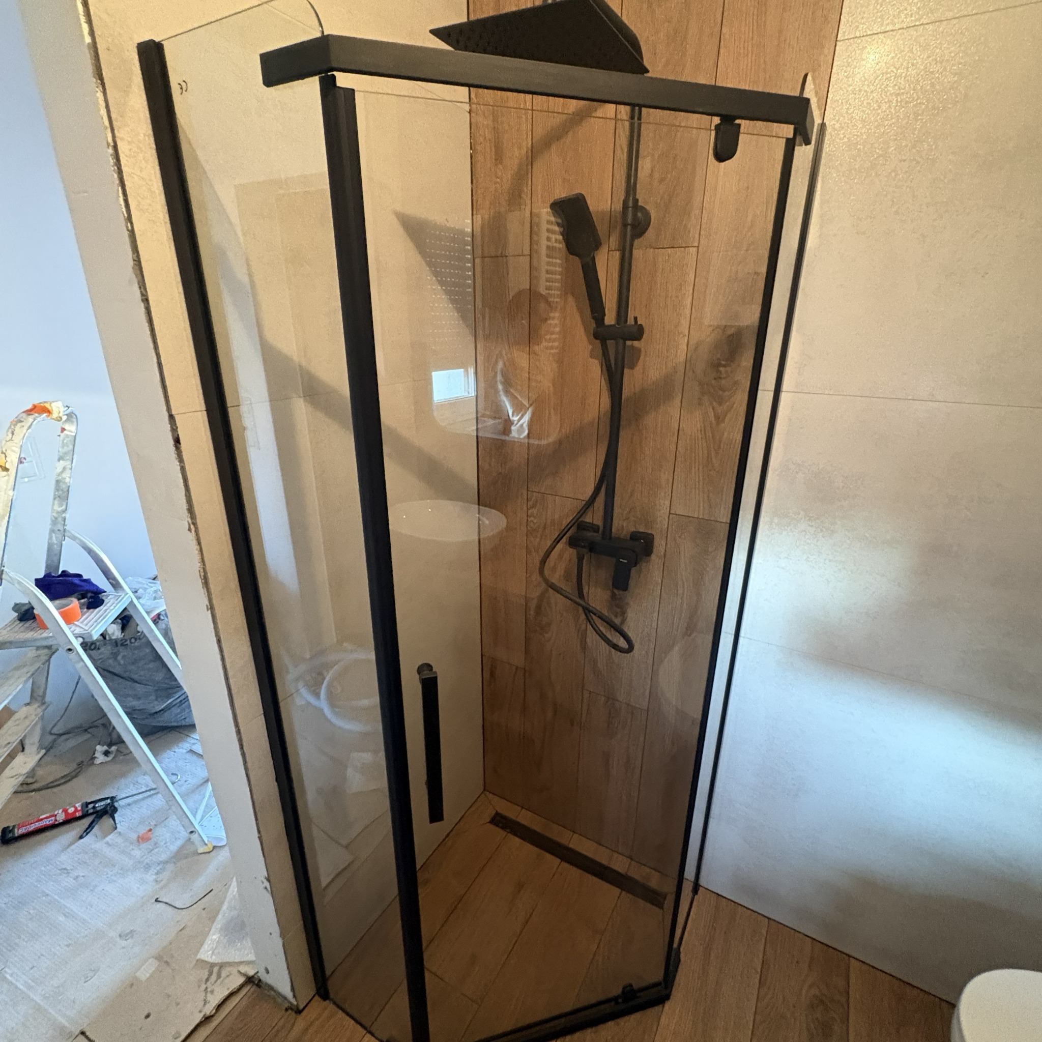 Kabina prysznicowa z czarnymi profilami i deszczownicą, wykończona płytkami imitującymi drewno. W tle drabina malarska i narzędzia.