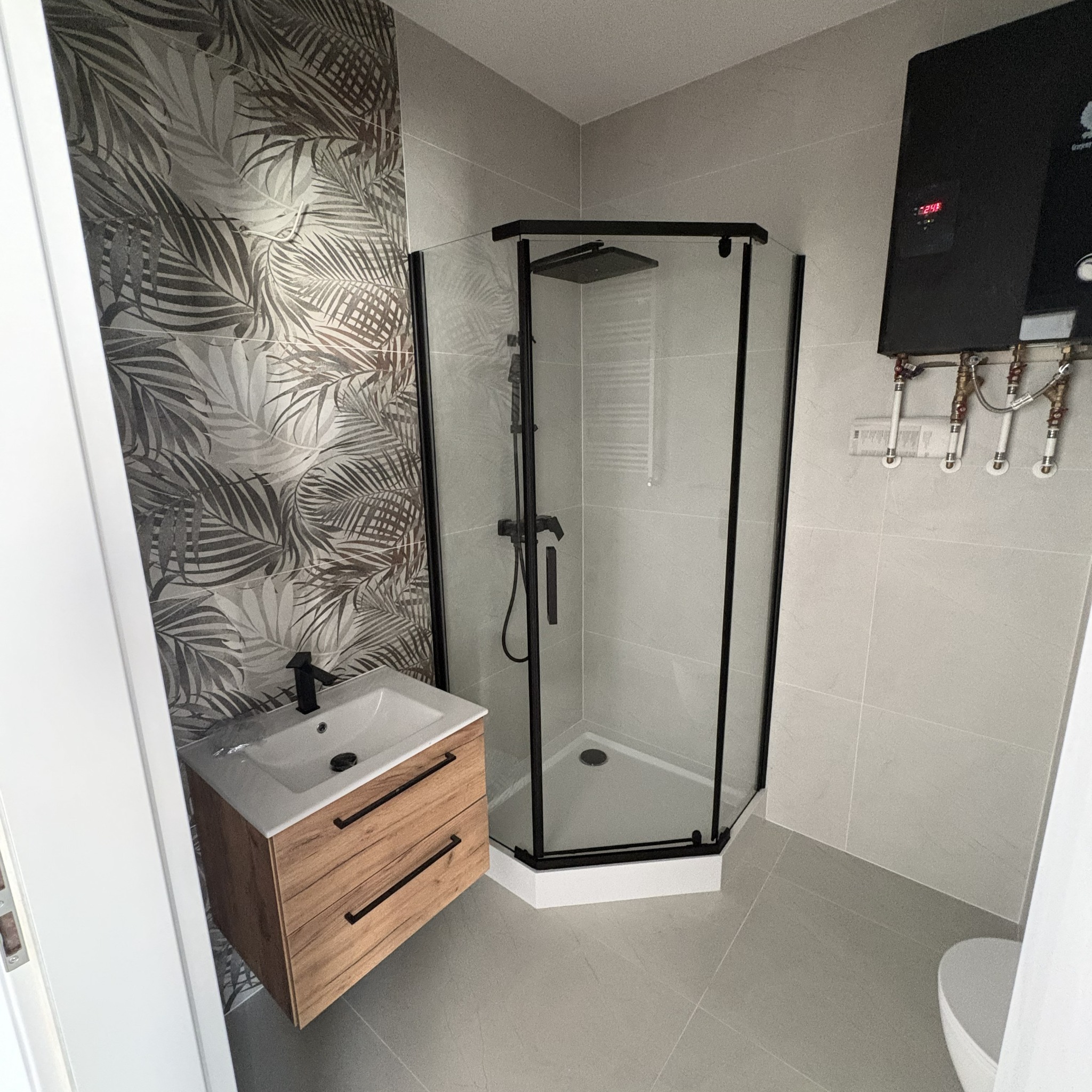 Wykończona łazienka z kabiną prysznicową w czarnej ramie, szafką pod umywalką z drewnianym frontem i płytkami z motywem liści. Widoczny bojler.