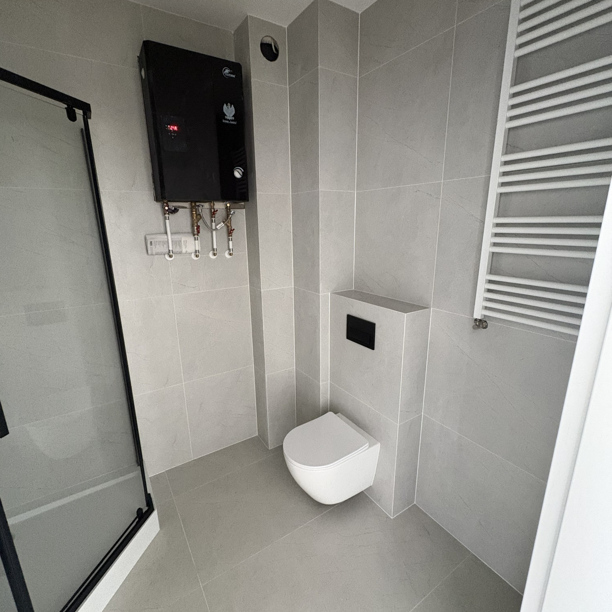 Nowoczesna łazienka w szarościach: wisząca toaleta, grzejnik drabinkowy i czarny bojler elektryczny. Minimalistyczny design, płytki imitujące kamień.