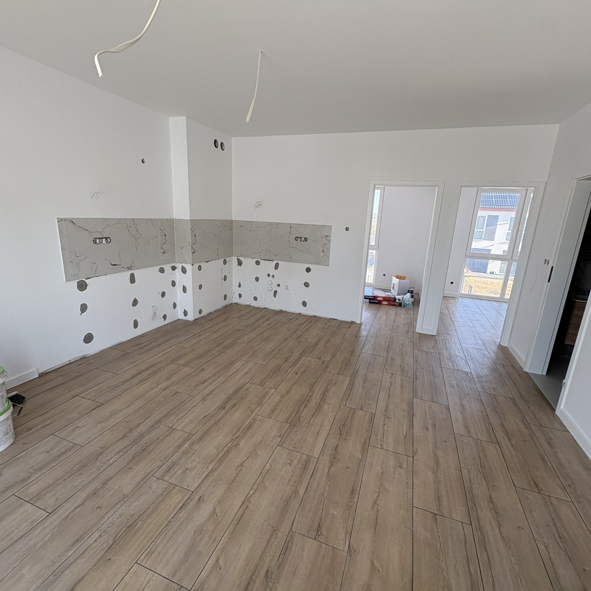 Wnętrze w trakcie wykańczania: drewnopodobna podłoga, fragment ściany z szarą płytką i otwory po gniazdkach, widoczne kable. Dwa otwarte przejścia do innych pomieszczeń.