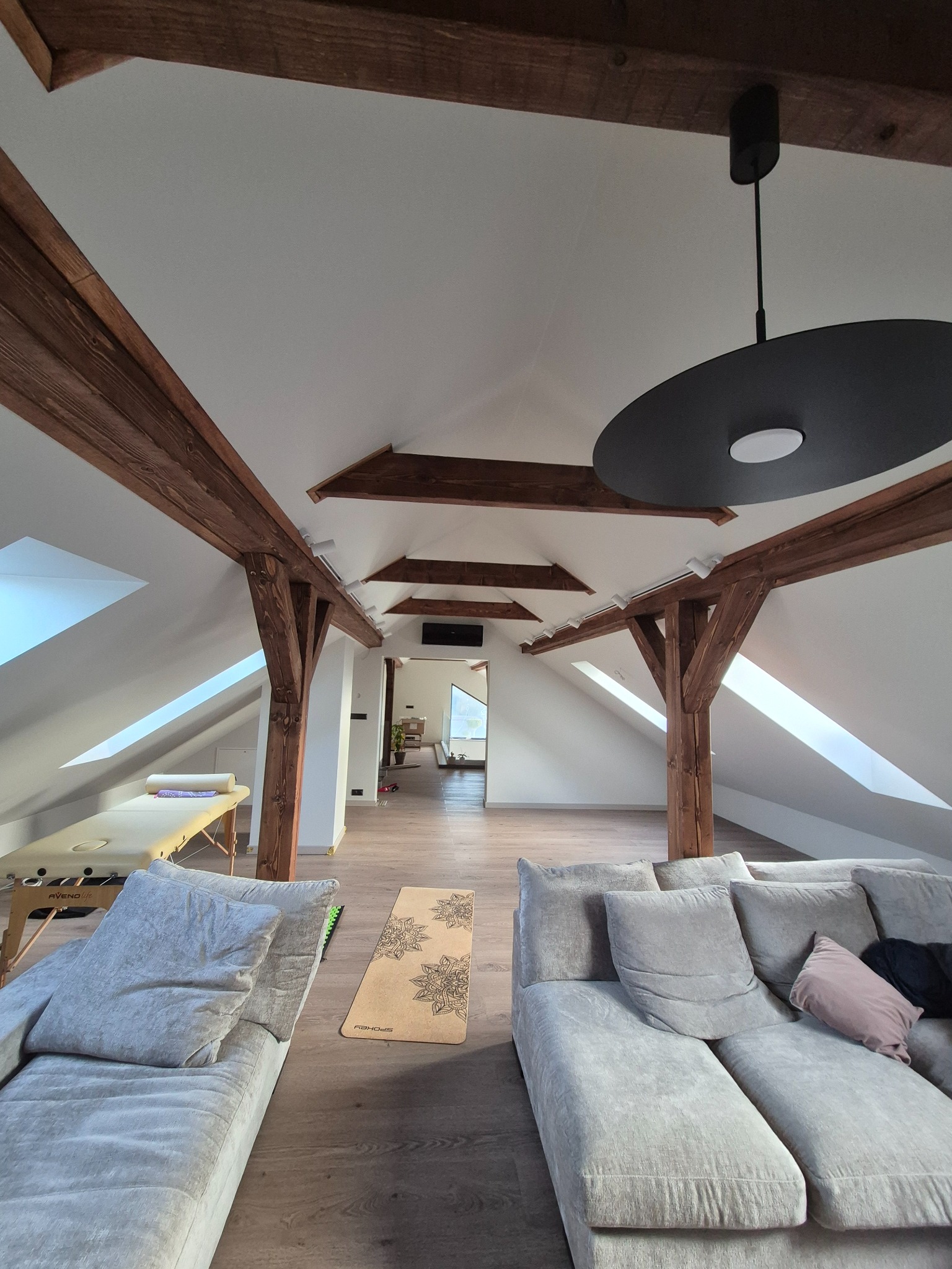 Jasne poddasze z drewnianymi belkami, szarą sofą, matą do ćwiczeń i kozetką. Widoczne skosy i okna dachowe. Minimalistyczny wystrój wnętrza.