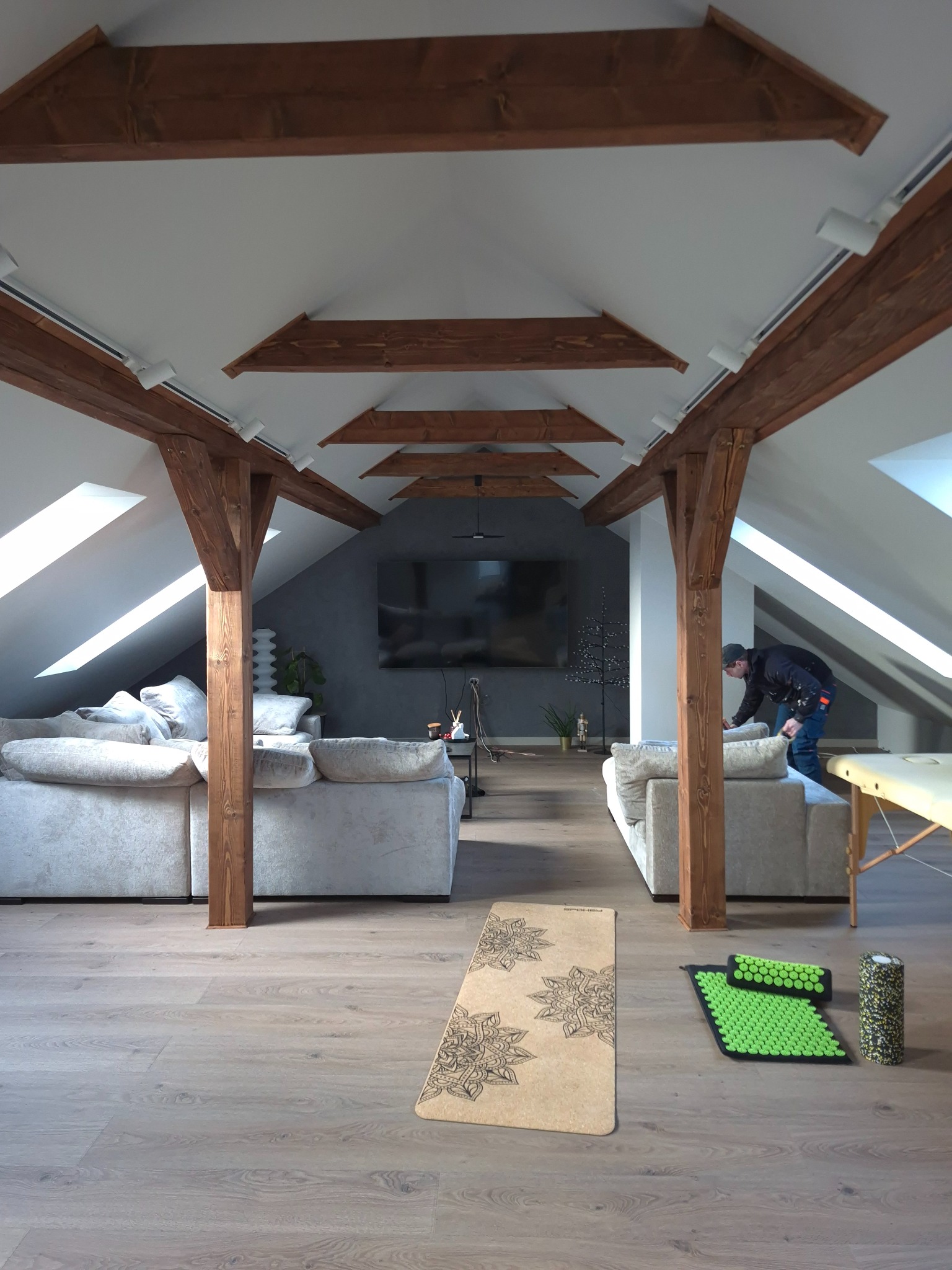 Jasne poddasze z drewnianymi belkami i oświetleniem punktowym. Widoczny fragment sofy, podłoga z paneli i akcesoria do ćwiczeń. Osoba w tle przy sofie.