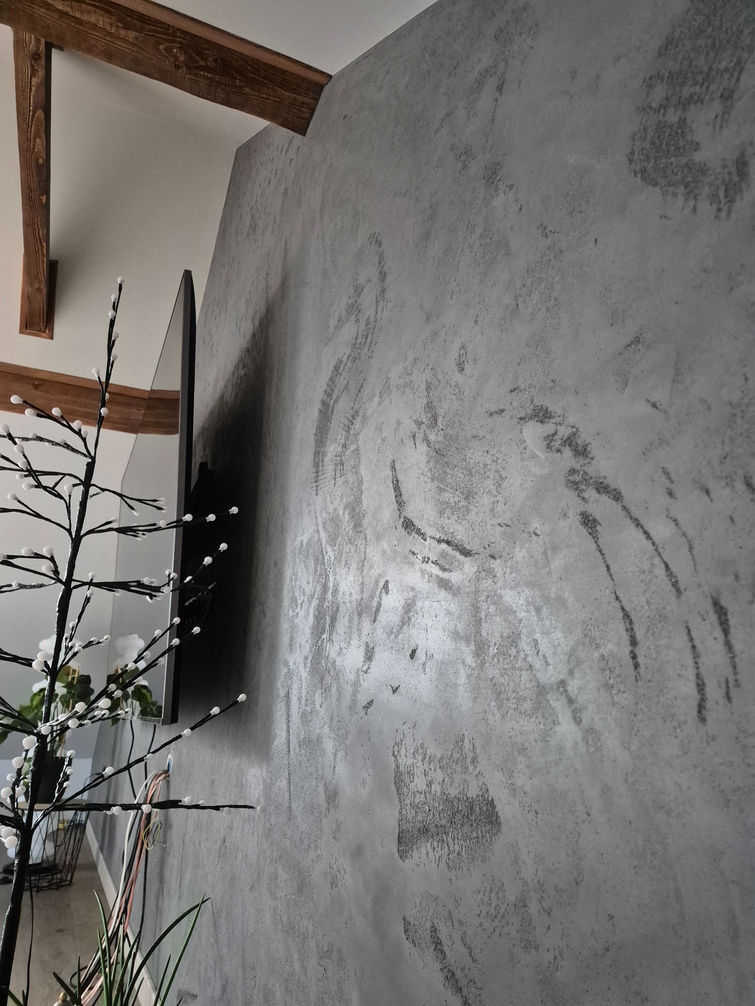 Szara ściana z efektem dekoracyjnym, widoczna z boku, z elementami drewnianymi na suficie i dekoracją roślinną w rogu. Widoczne przewody elektryczne.