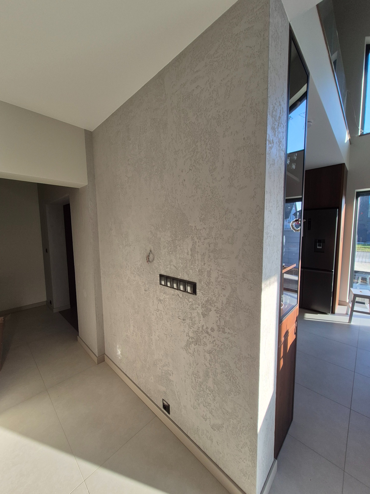 Ściana z dekoracyjnym tynkiem w odcieniach szarości, widoczne gniazdka elektryczne. W tle fragment kuchni z zabudową i oknem. Jasne płytki na podłodze.