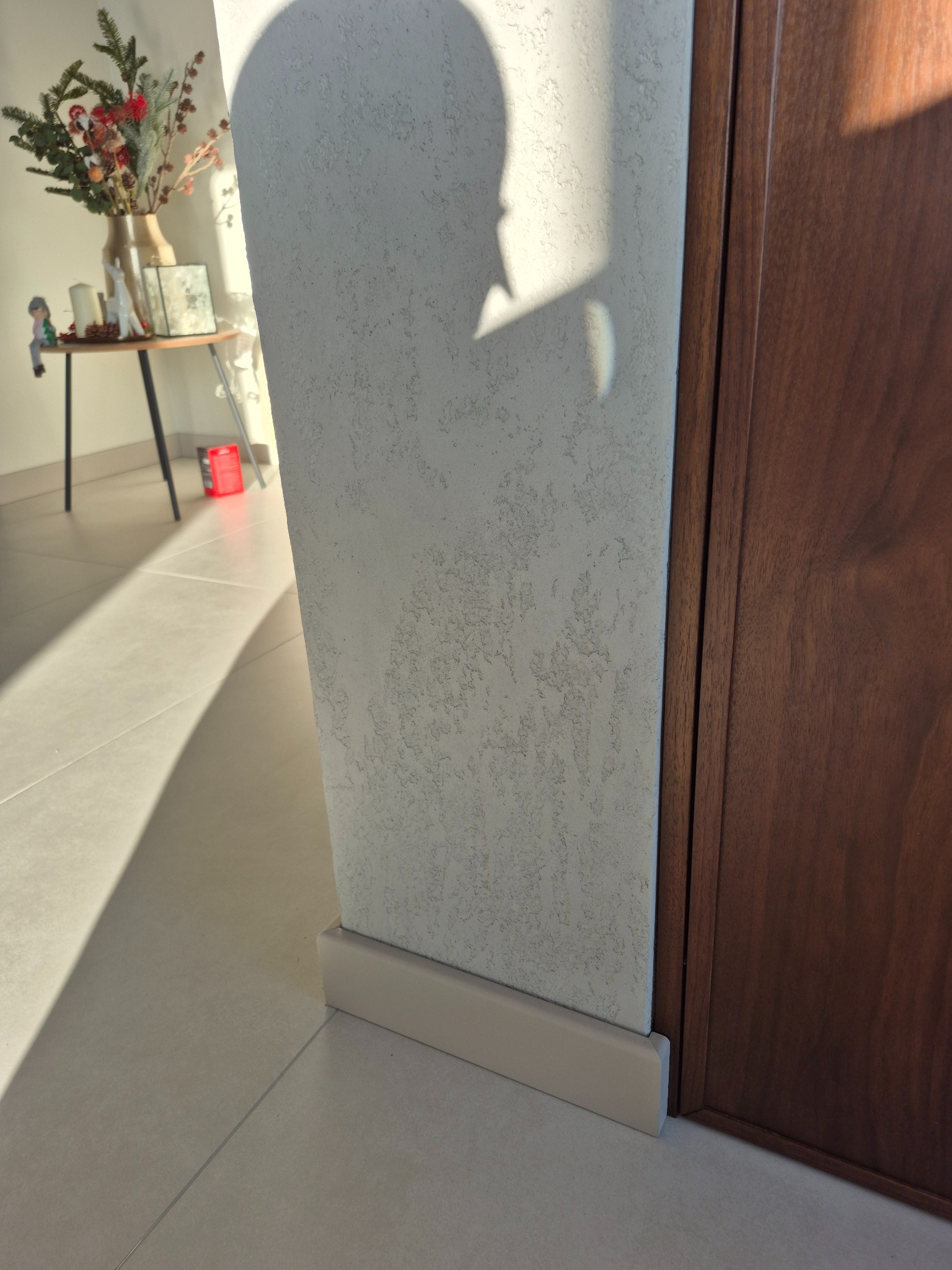Fragment ściany z fakturą, listwa przypodłogowa i fragment drewnianych drzwi. Widoczne światło słoneczne i cień. W tle stolik z dekoracjami.