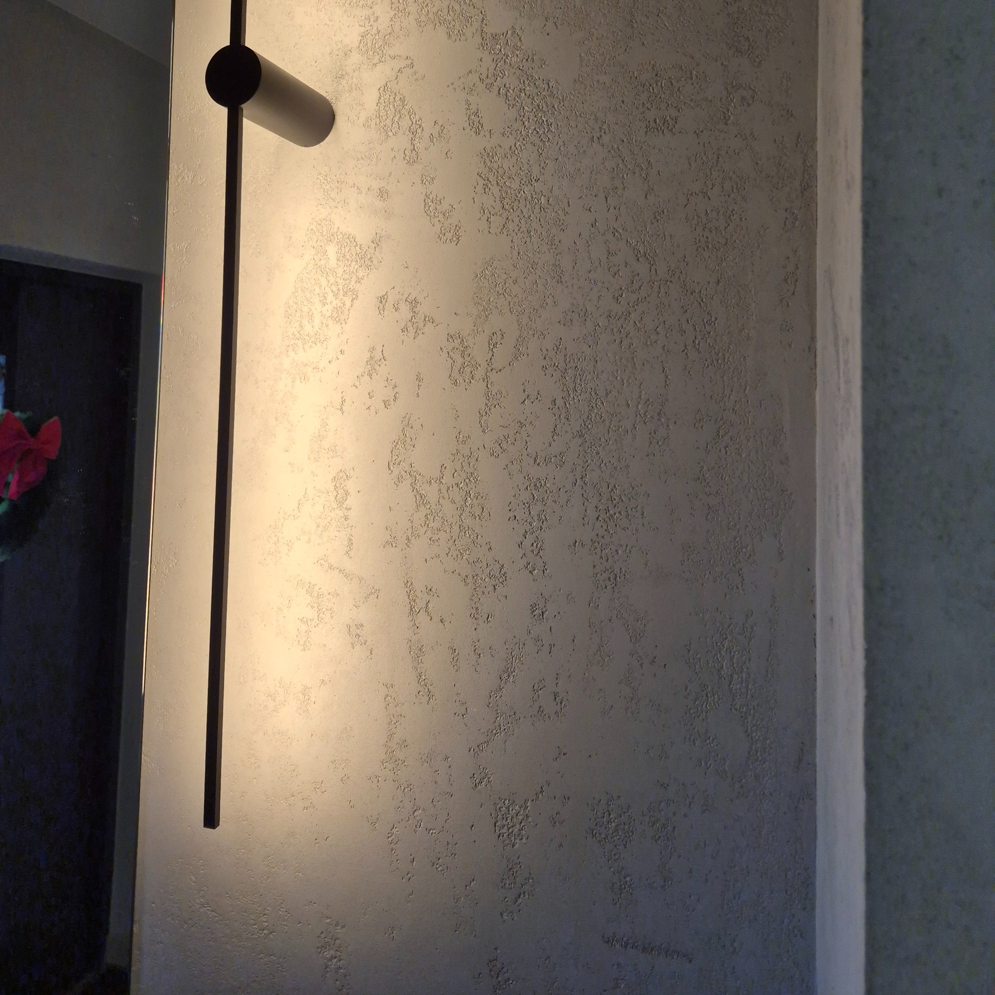 Szara ściana z dekoracyjną fakturą i nowoczesnym oświetleniem punktowym. Widoczny fragment lustra z odbiciem. Minimalistyczny design wnętrza.