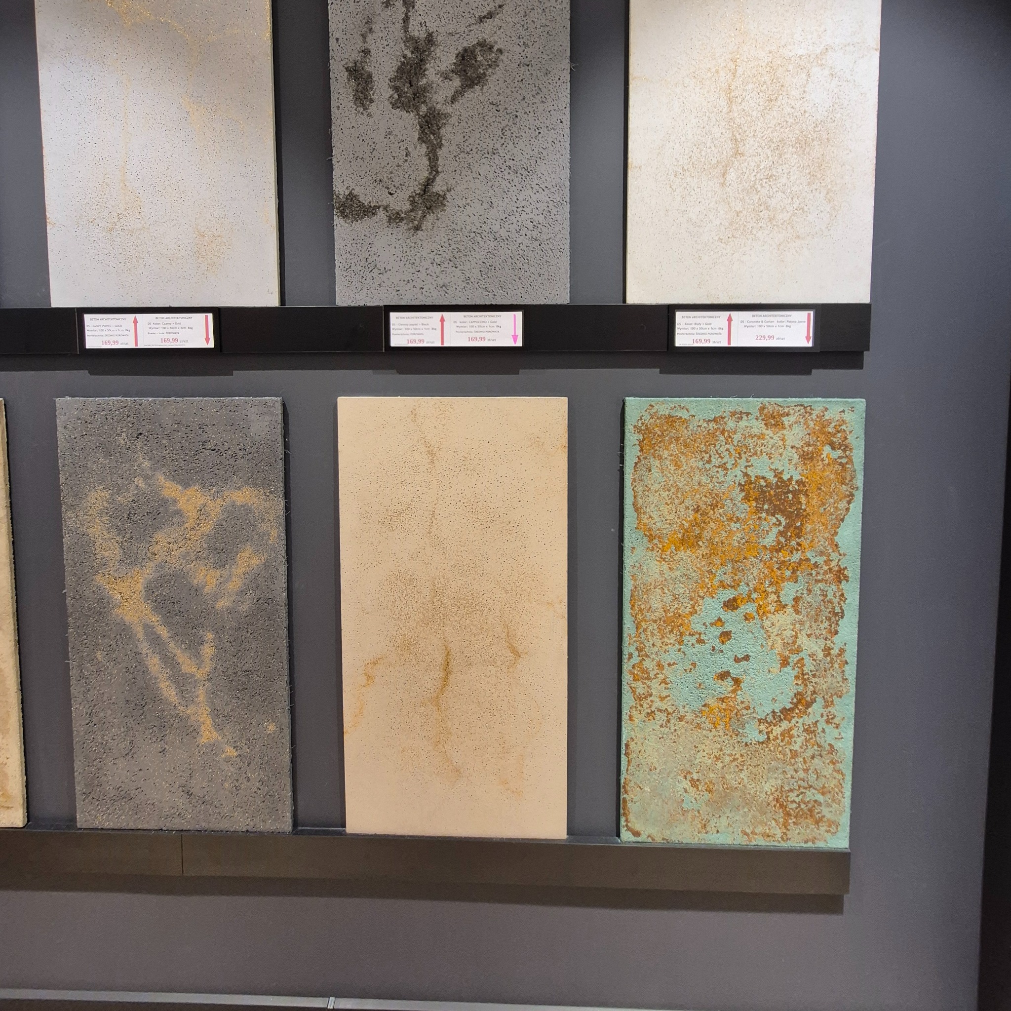 Ekspozycja paneli dekoracyjnych imitujących beton architektoniczny w różnych kolorach i fakturach, z widocznymi cenami i specyfikacją techniczną.