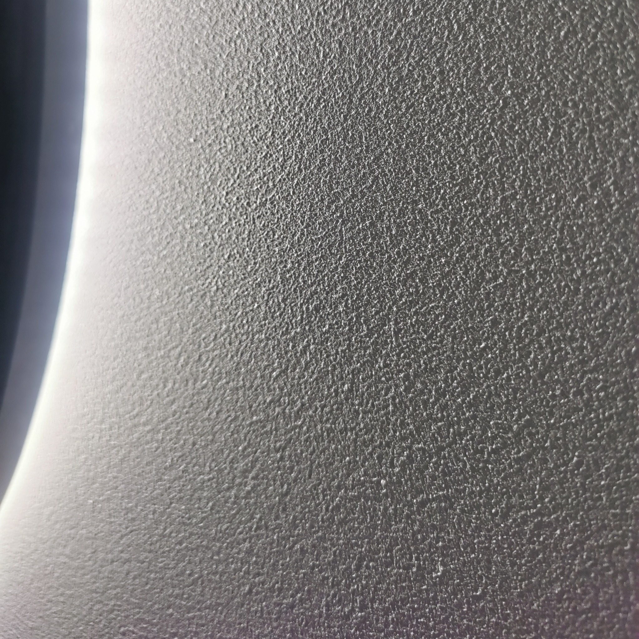 Szara, teksturowana powierzchnia ściany z widoczną strukturą, oświetlona z lewej strony, tworząca gradient światła i cienia. Delikatna faktura tynku dekoracyjnego.