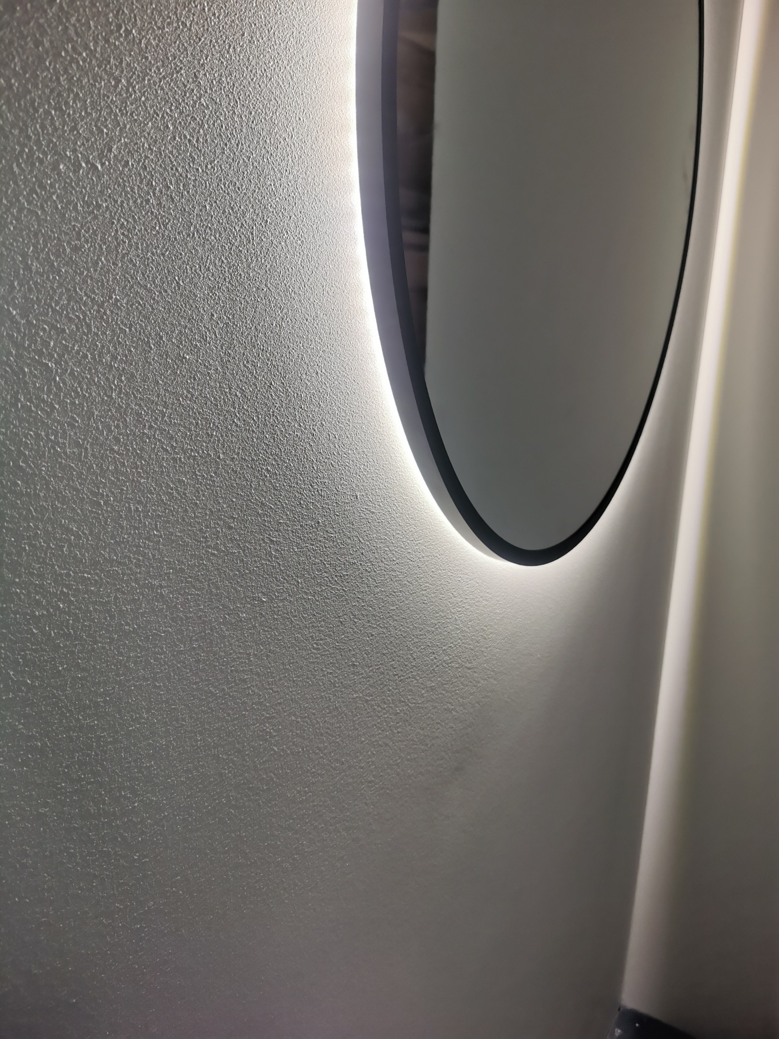 Owalne lustro z podświetleniem LED na teksturowanej, jasnej ścianie. Minimalistyczny design, nowoczesne wykończenie. Oświetlenie akcentujące.