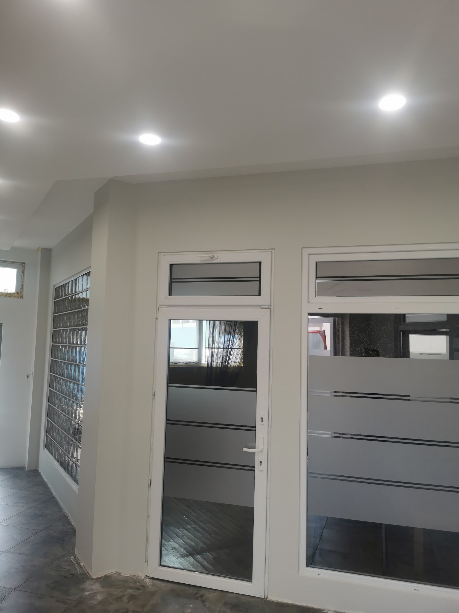 Białe drzwi z poziomymi pasami folii dekoracyjnej, szklana ściana z luksferów, świeżo pomalowane ściany i sufit z oświetleniem punktowym. Nowoczesne wykończenie.