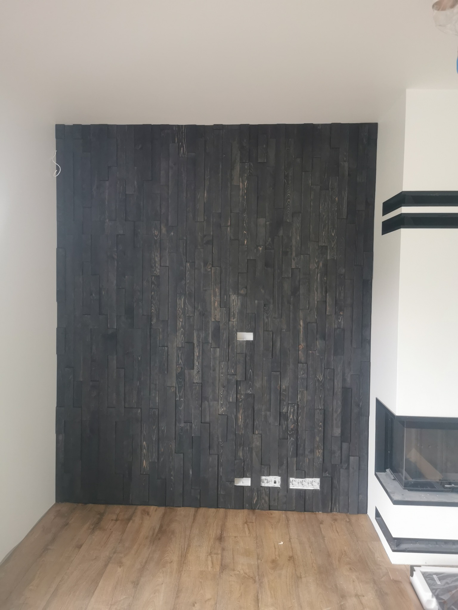 Ściana z ciemnego drewna ułożonego w pionowe pasy, z widocznymi gniazdkami elektrycznymi i fragmentem kominka w nowoczesnym wnętrzu z drewnianą podłogą.