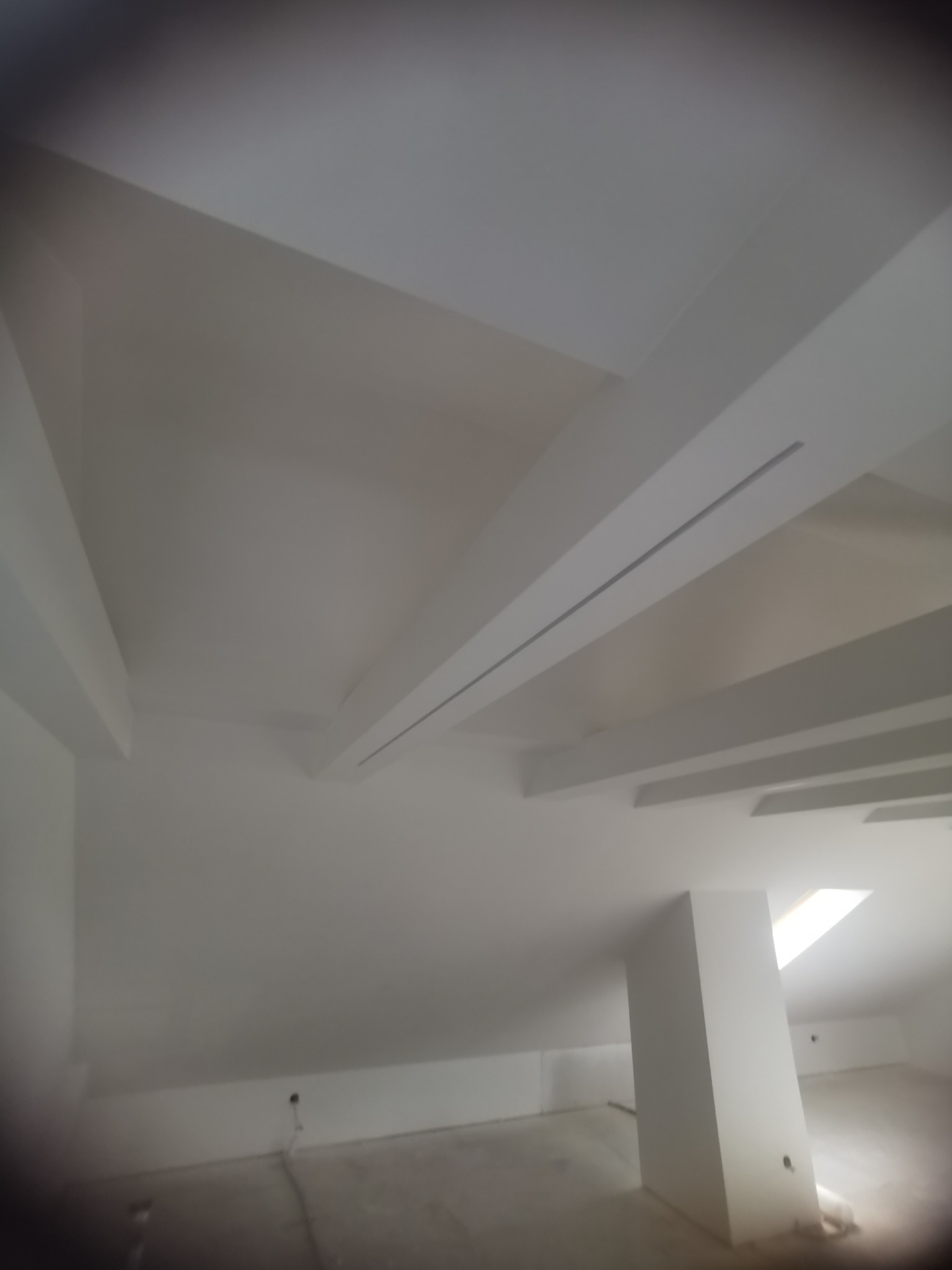 Białe, świeżo pomalowane poddasze ze skosami i belkami stropowymi. Widoczne okno dachowe i kolumna nośna. Minimalistyczny design, surowe wykończenie.