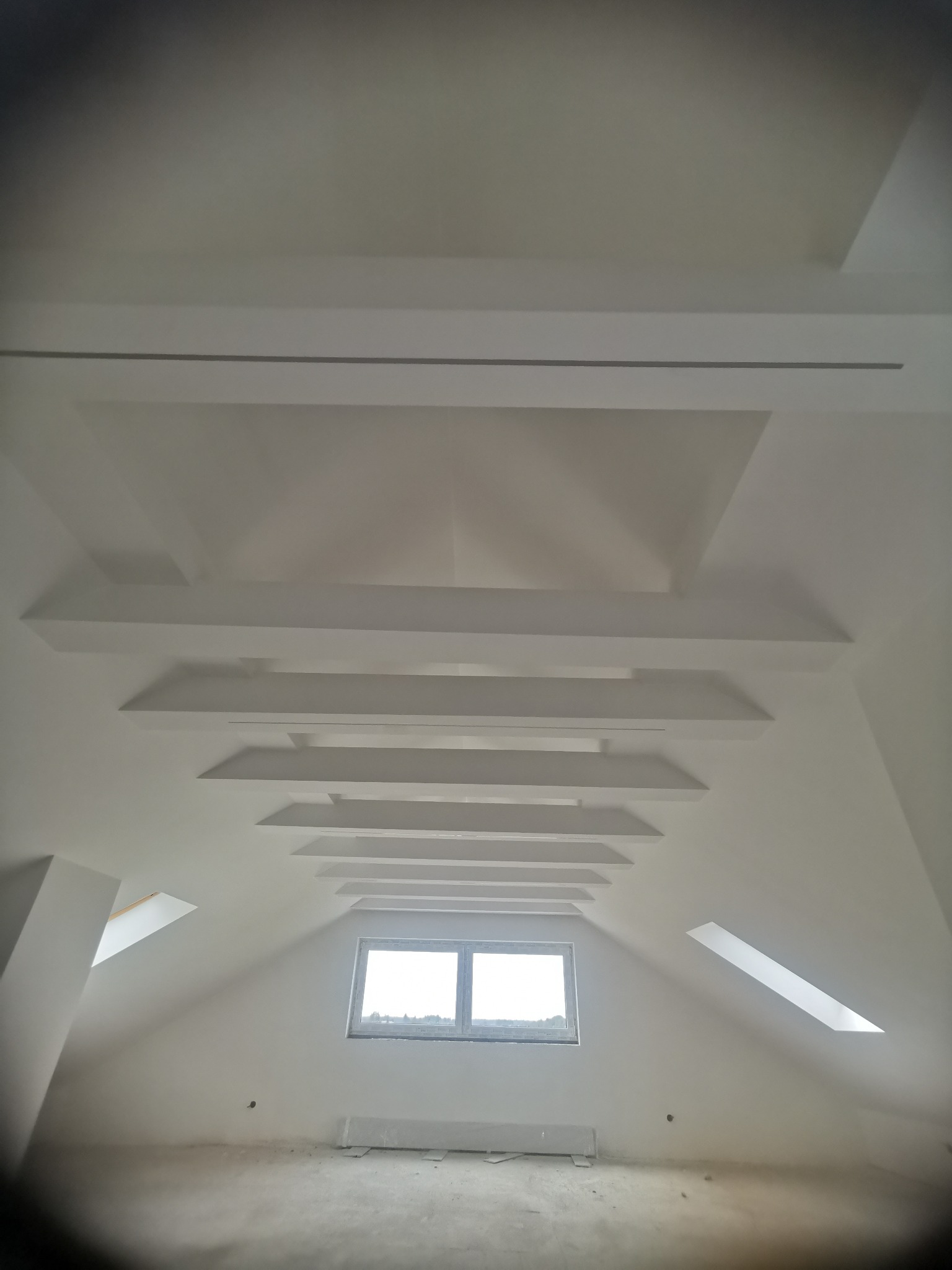 Białe, symetryczne belki na poddaszu z oknem dachowym. Widok z dołu, podkreślający geometrię sufitu i skosy ścian. Minimalistyczny design, surowy stan wykończenia.