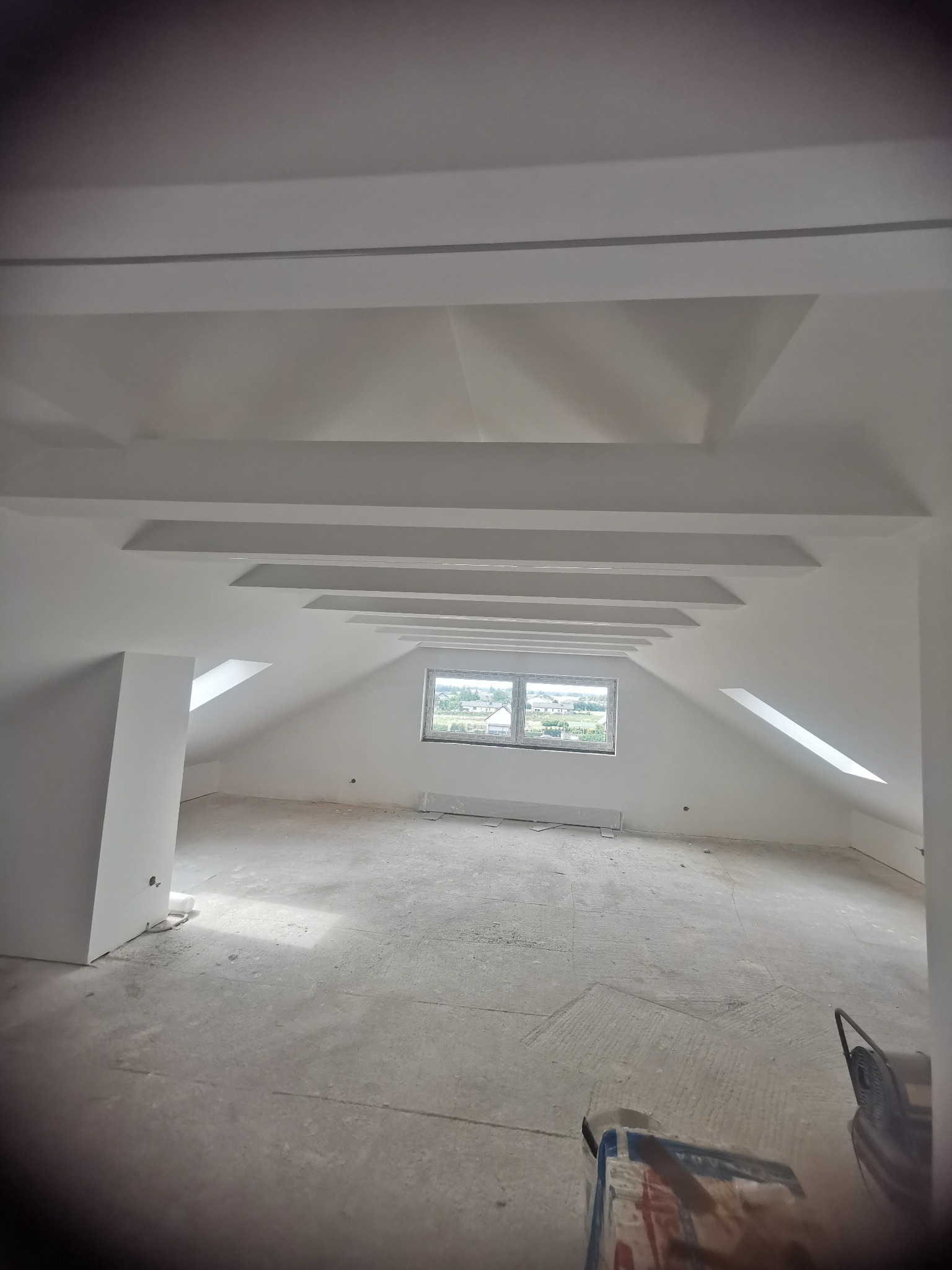 Wykończone poddasze ze skosami i oknem w Tarnowskich Górach. Sufit z dekoracyjnymi elementami gipsowymi. Ściany i sufit pomalowane na biało. Podłoga surowa, przygotowana do dalszych prac.