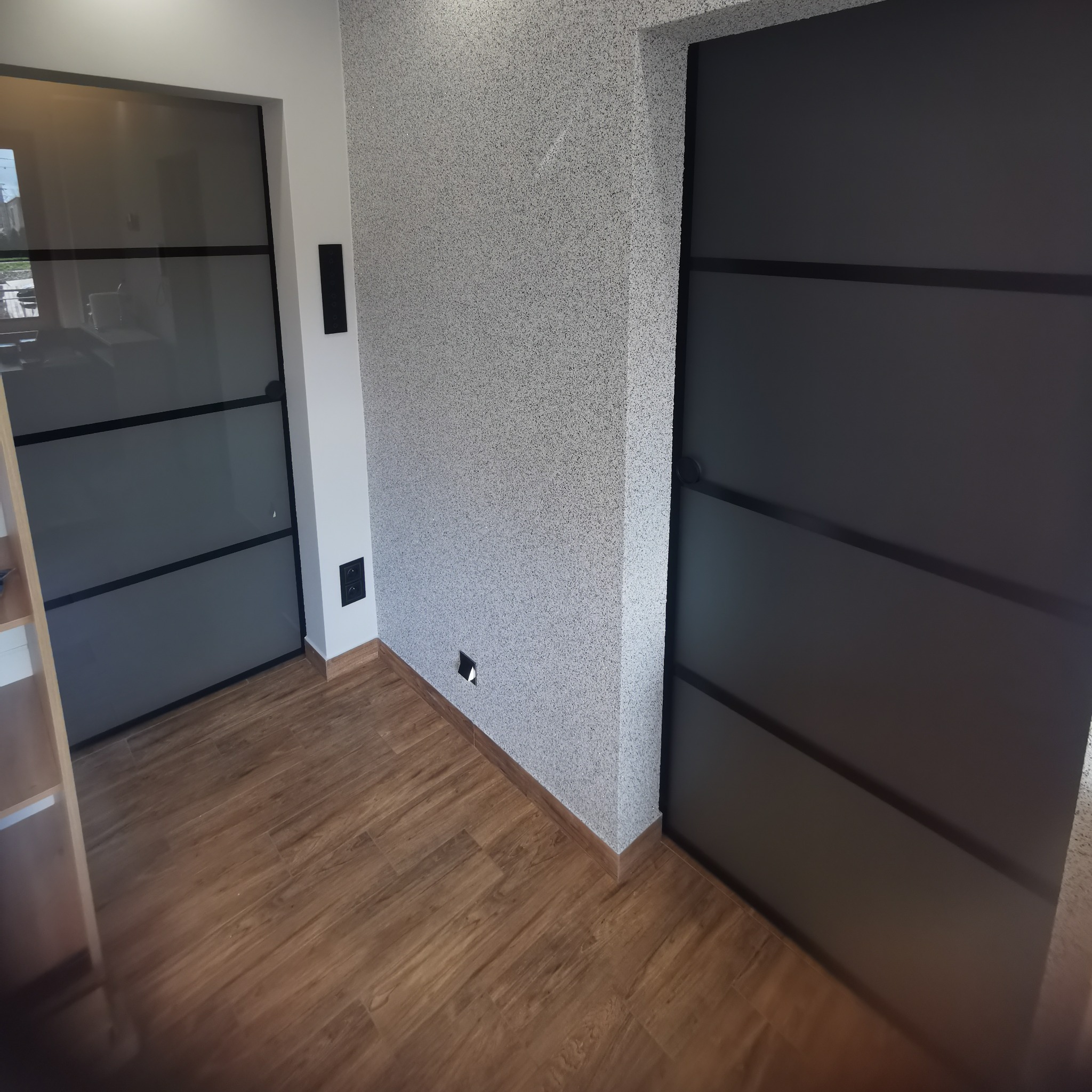 Nowoczesne wnętrze z drewnianą podłogą, ścianą o fakturze baranka i dwoma parami drzwi z czarnymi ramami, z czego jedne są przeszklone.