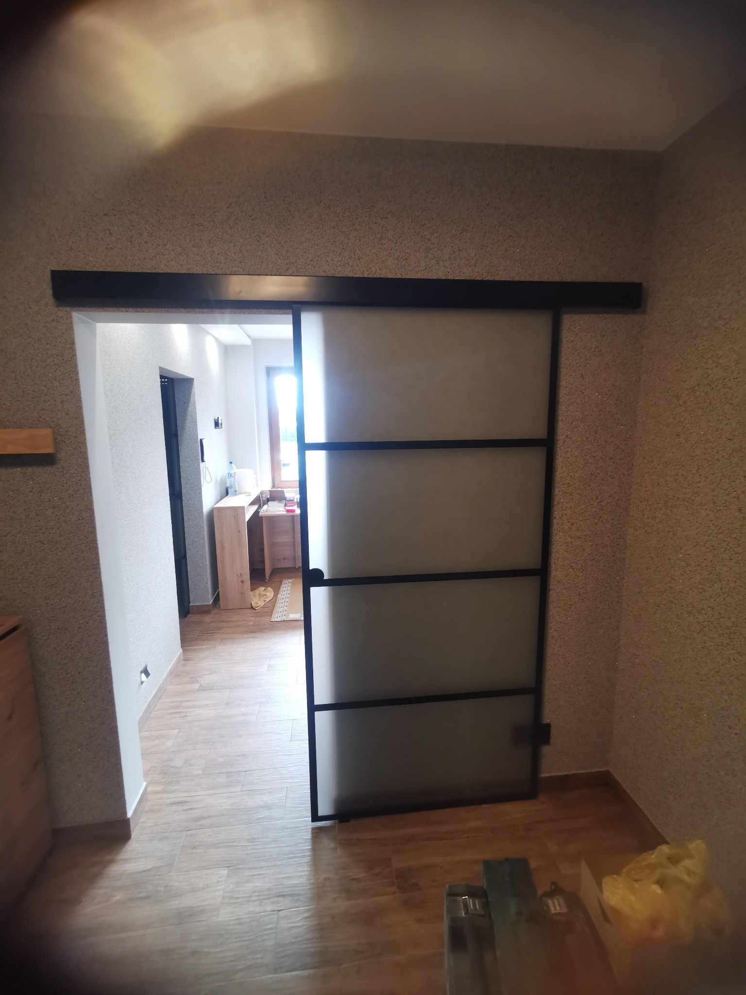 Nowoczesne drzwi przesuwne z czarną ramą i matowym szkłem, zamontowane w przejściu między pomieszczeniami. Widoczne narzędzia i materiały montażowe.