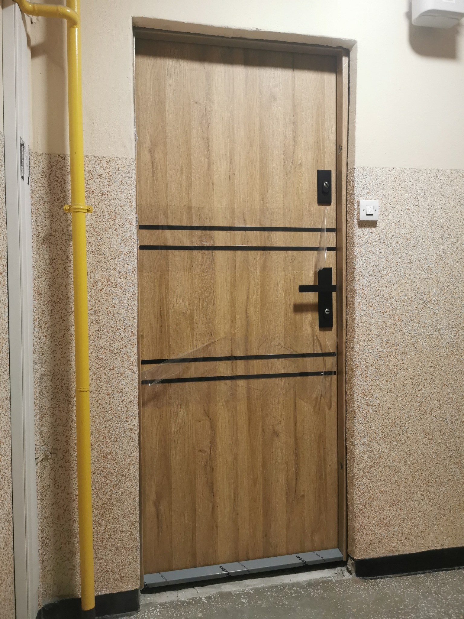 Zamontowane drzwi wewnętrzne z drewnianym wykończeniem, zabezpieczone folią ochronną, widoczne na tle ściany z charakterystyczną fakturą i żółtą rurą gazową.