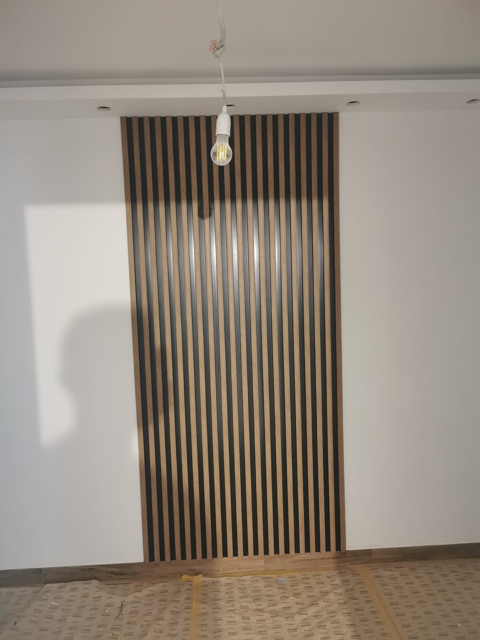 Ściana z dekoracyjnymi panelami lamelowymi w odcieniach drewna i czerni, oświetlona wiszącą żarówką. Podłoga zabezpieczona folią ochronną.