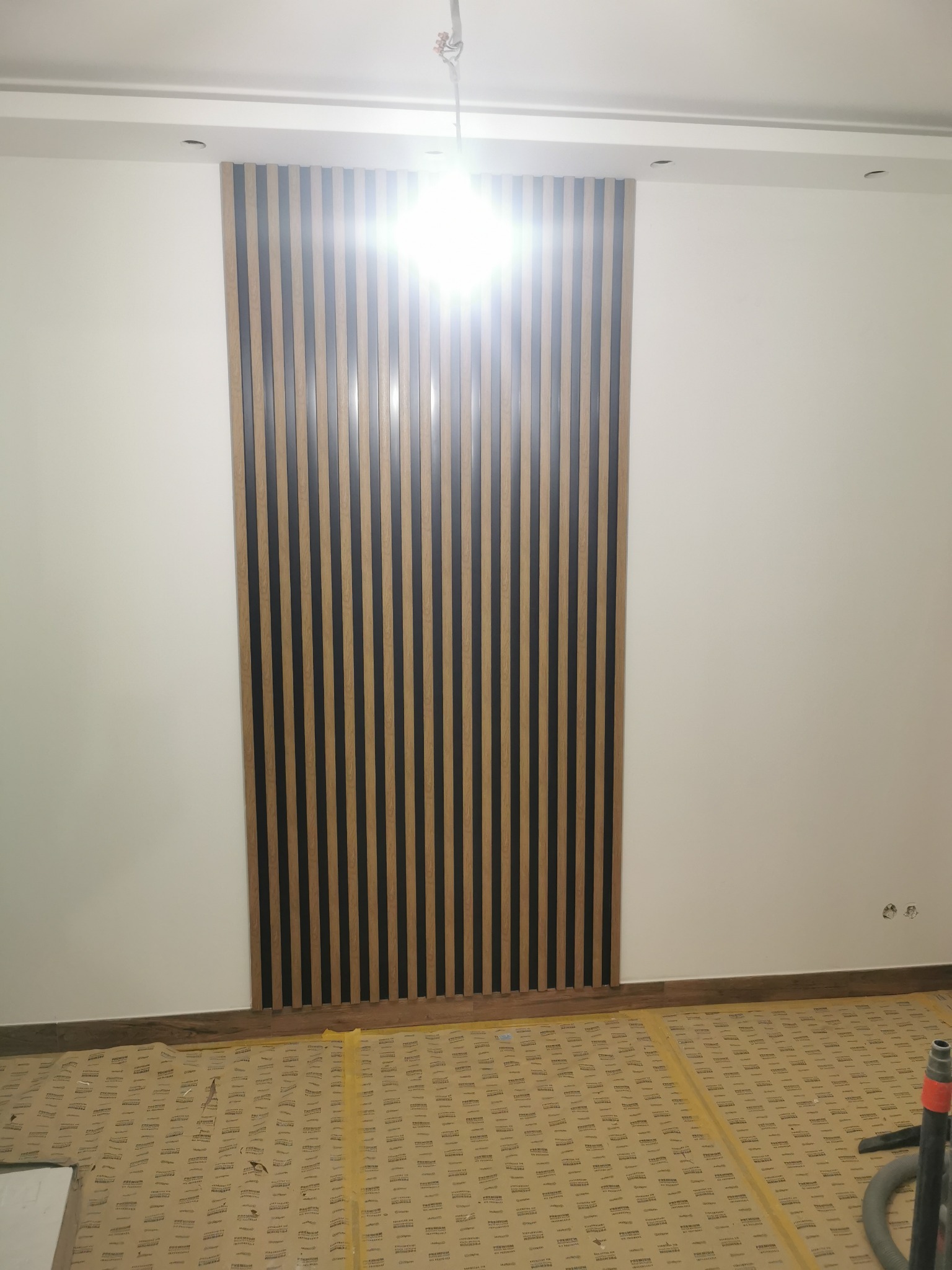 Ściana z dekoracyjnymi panelami pionowymi w odcieniach drewna i czerni. Podłoga zabezpieczona kartonem. Widoczne gniazdka elektryczne.