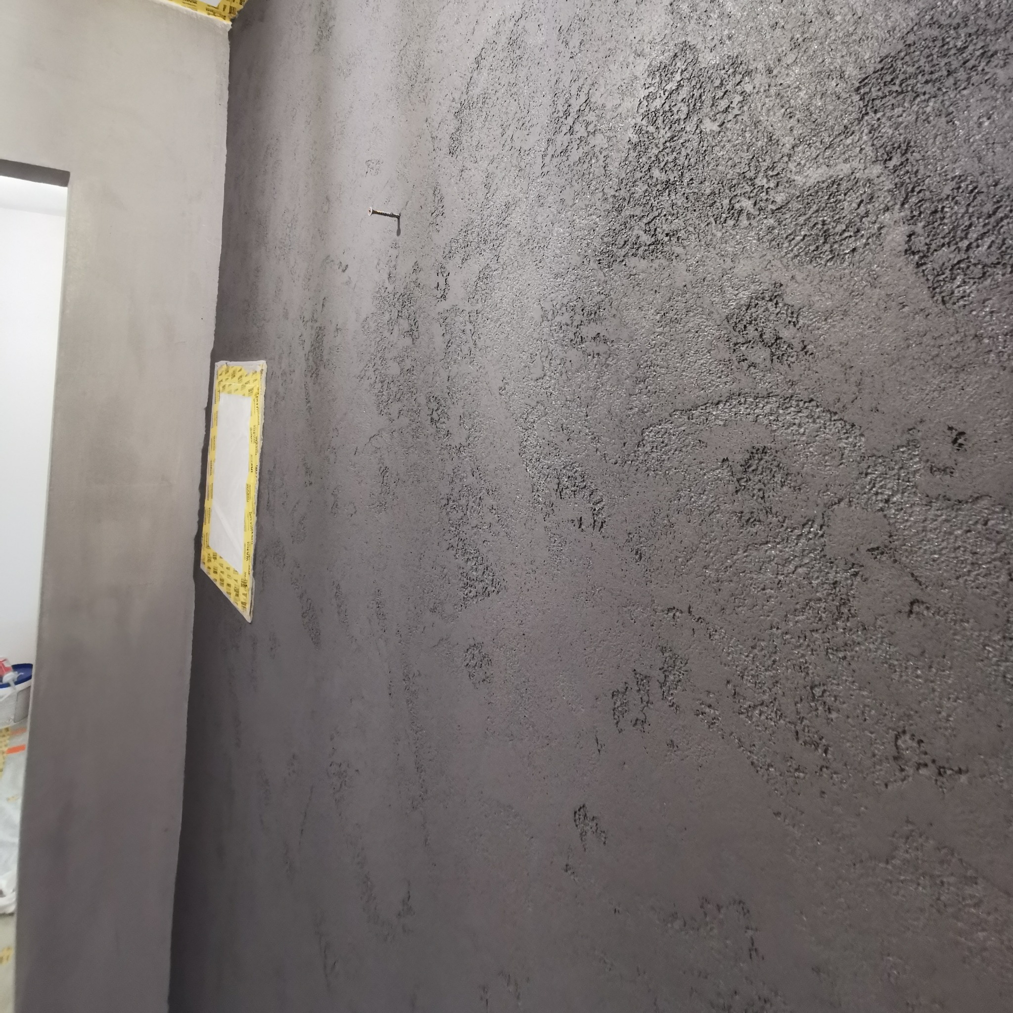 Ściana z szarą, dekoracyjną strukturą, zabezpieczona taśmą malarską wokół otworu. Widoczny fragment drugiej ściany w surowym stanie.