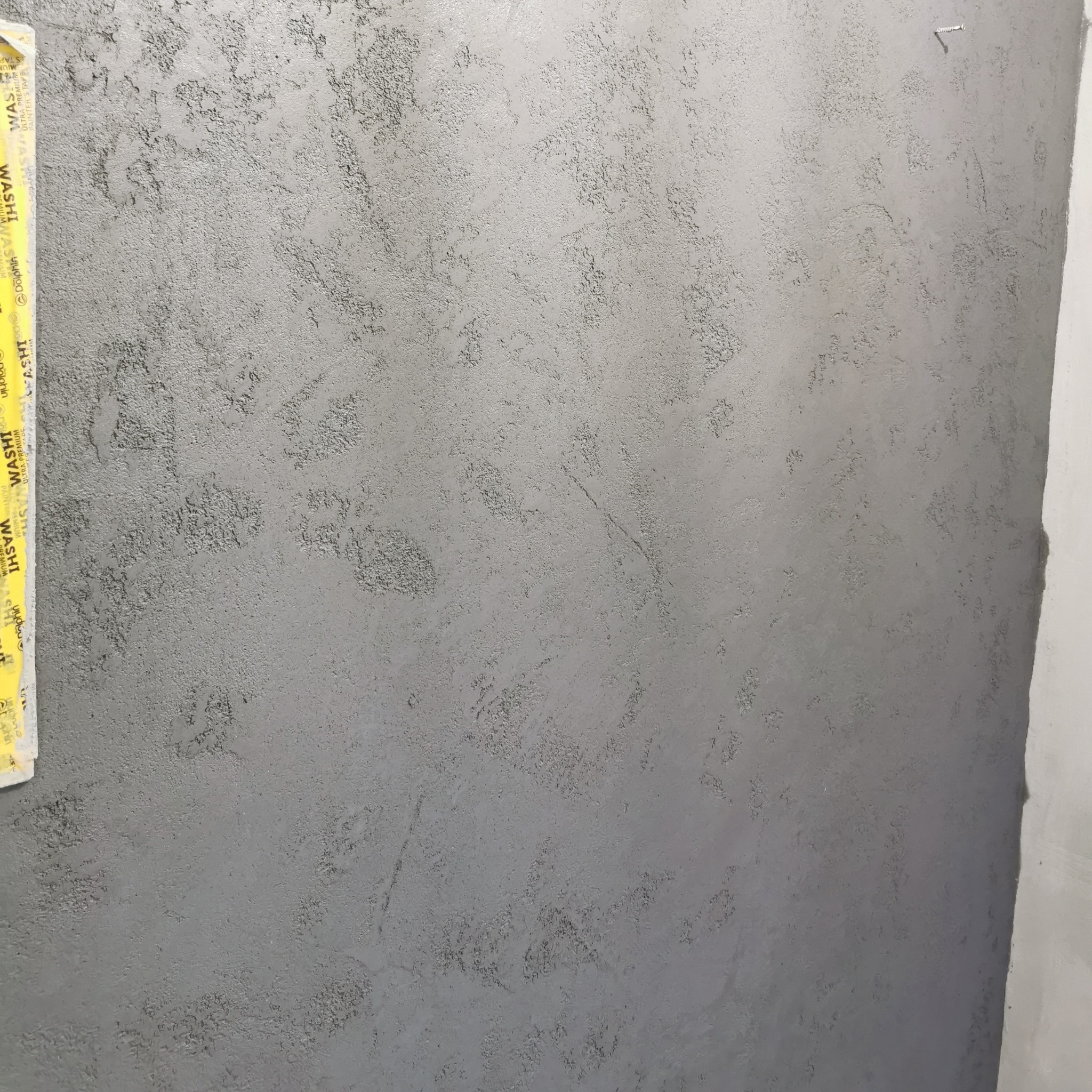 Szara ściana z dekoracyjną strukturą, widoczna taśma malarska przy krawędzi. Delikatna faktura i neutralna kolorystyka. Minimalistyczny detal wykończenia.