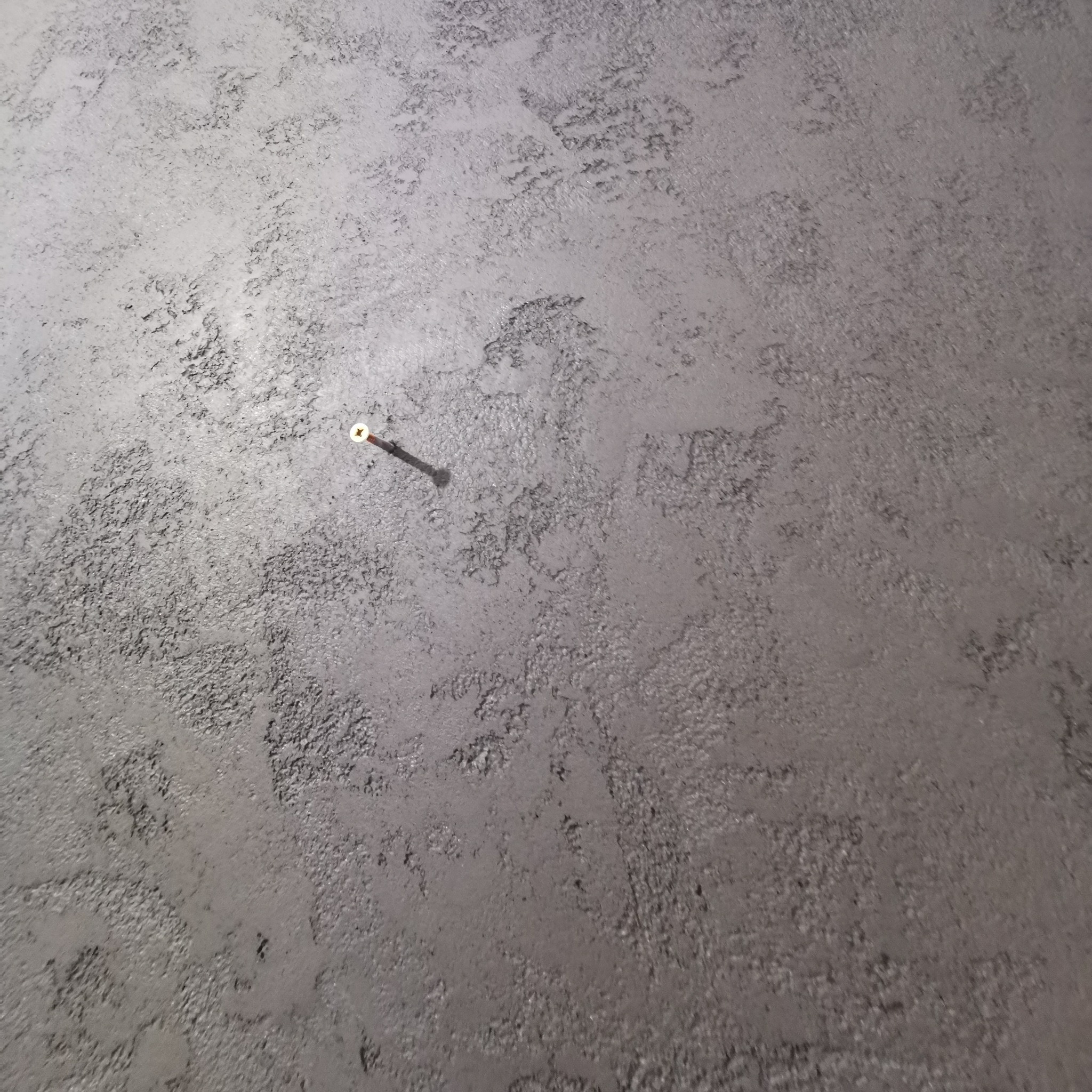 Szara, teksturowana ściana z widocznym gwoździem. Delikatna faktura i stonowana kolorystyka. Minimalistyczny detal wykończenia.