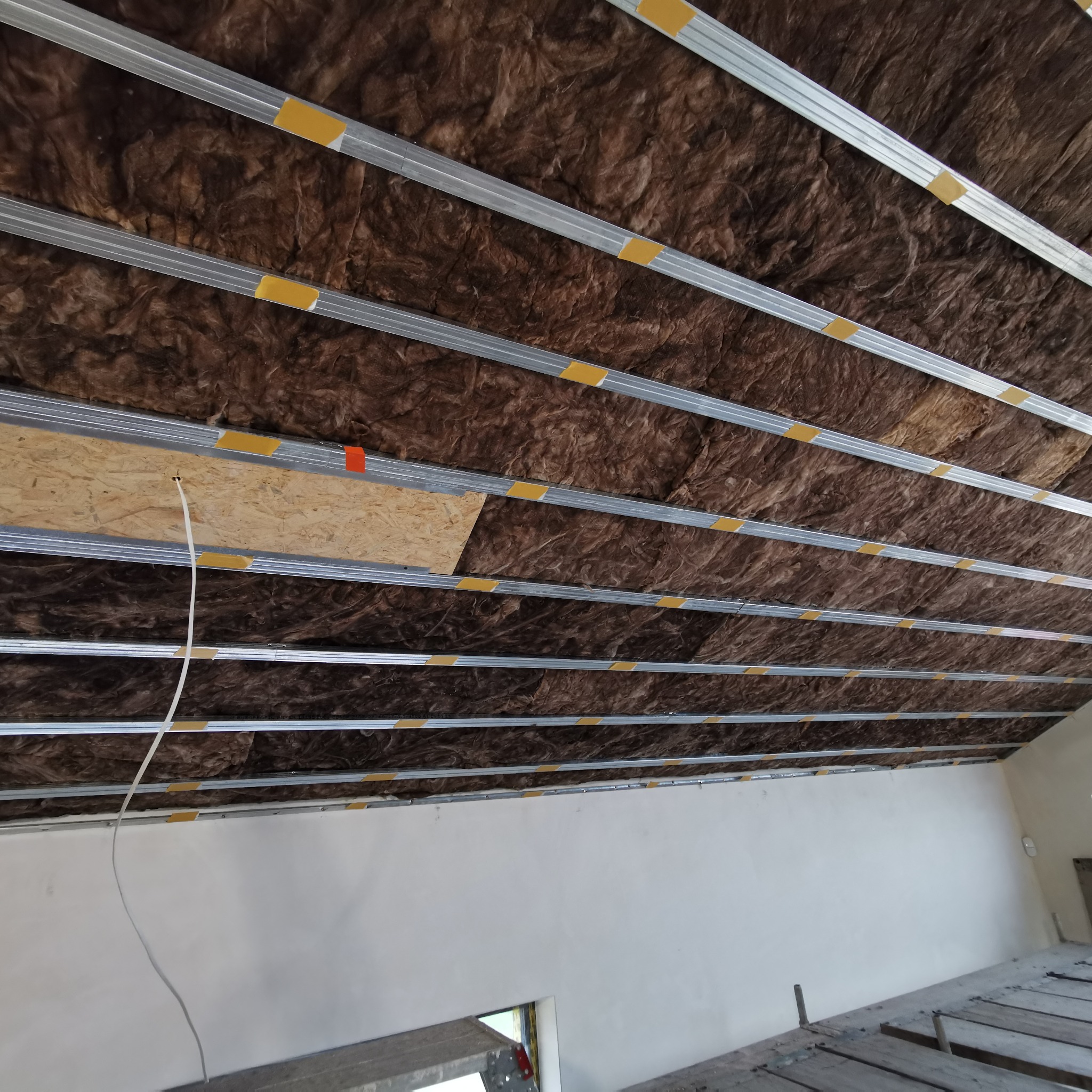 Sufit podwieszany w trakcie montażu: metalowa konstrukcja, izolacja z wełny mineralnej, fragment płyty OSB z kablem. Widok z dołu na sufit w trakcie prac.