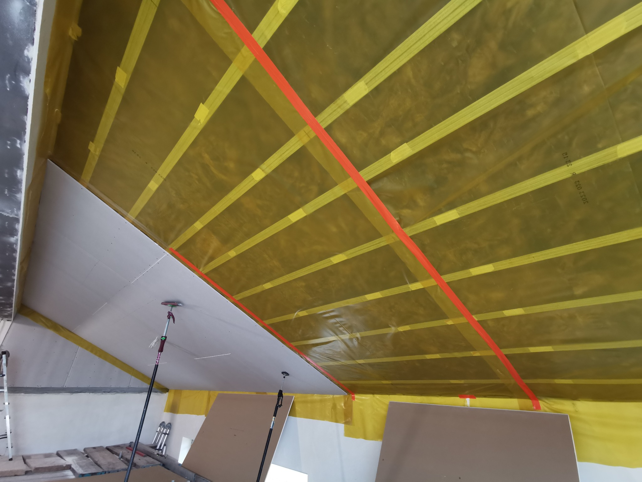 Skośny sufit poddasza w trakcie prac: widoczne płyty, folia paroizolacyjna z łatami i czerwona taśma. Użycie podpór teleskopowych. Etap przed szpachlowaniem.