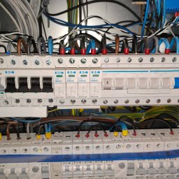 Rozdzielnica elektryczna z widocznymi bezpiecznikami, przewodami w różnych kolorach i oznaczeniami. Widok z bliska na instalację elektryczną wewnątrz skrzynki.