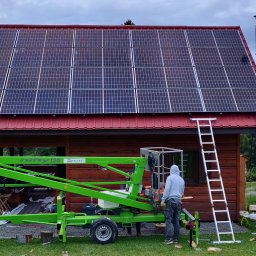 Gogik.pl Sp. z o.o. - Montaż paneli słonecznych na dachu drewnianego domu z użyciem podnośnika. Widoczne drabiny i osoba w bluzie z kapturem. W tle górzysty krajobraz.