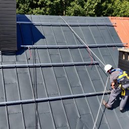 Gogik.pl Sp. z o.o. - Montaż paneli słonecznych na szarym dachu z blachy. Widoczny pracownik w kasku i uprzęży, częściowo zamontowany panel i szyny montażowe. W tle komin i drzewa.