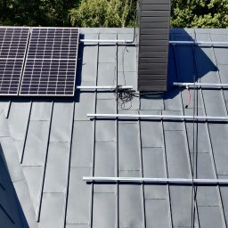 Gogik.pl Sp. z o.o. - Montaż paneli fotowoltaicznych na szarym dachu blaszanym, widok z góry. Częściowo zamontowane panele i szyny montażowe. W tle zieleń drzew.