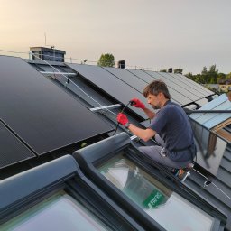 Gogik.pl Sp. z o.o. - Montaż paneli fotowoltaicznych na dachu z oknami dachowymi Fakro. Instalator w czerwonych rękawicach łączy przewody. Widok z góry, w tle zabudowa jednorodzinna.