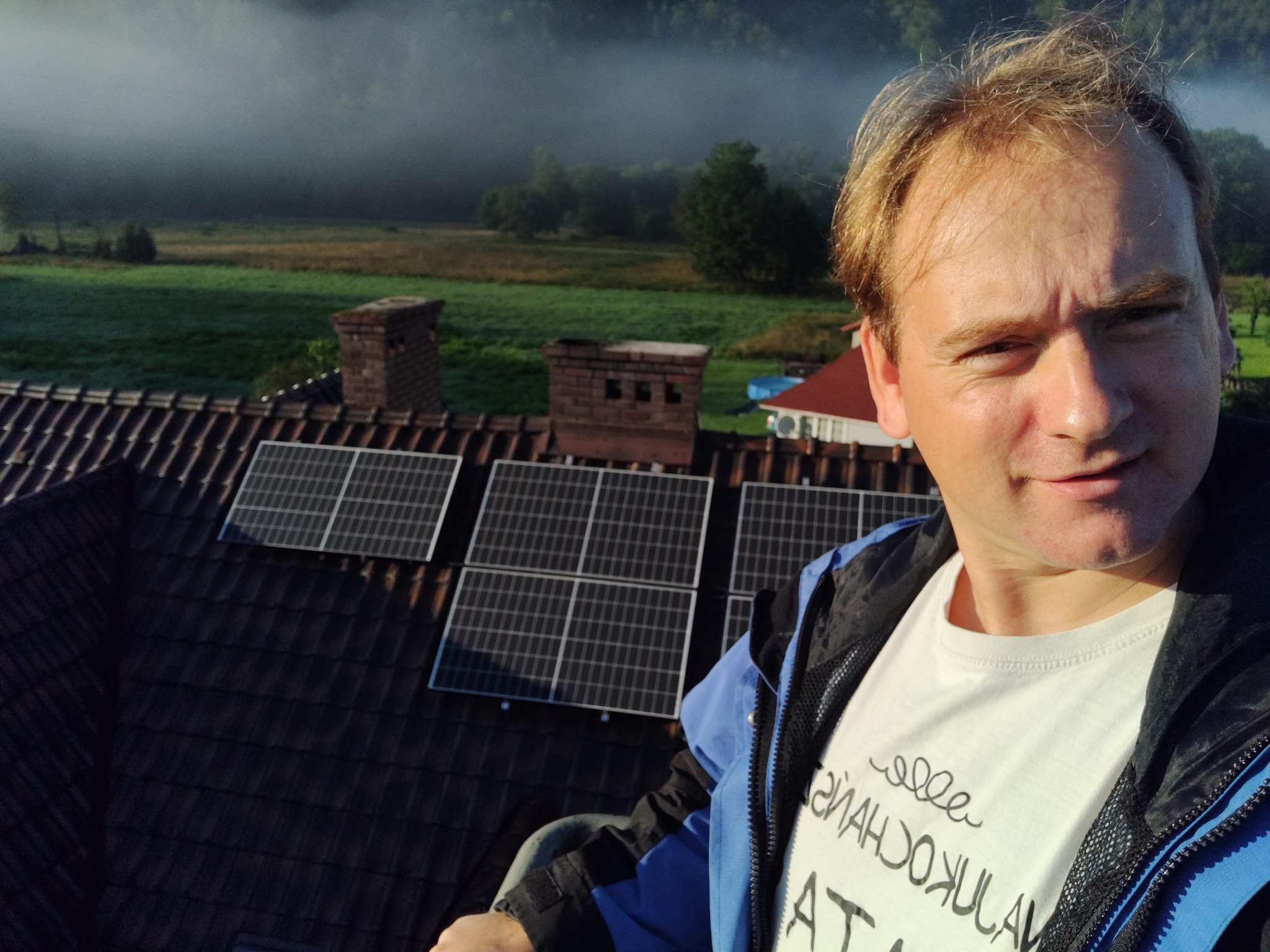 Instalacja paneli słonecznych na dachu z cegły, widok z góry, w tle zalesione wzgórza spowite mgłą. Osoba w kadrze, ubrana w kurtkę, patrzy w obiektyw.