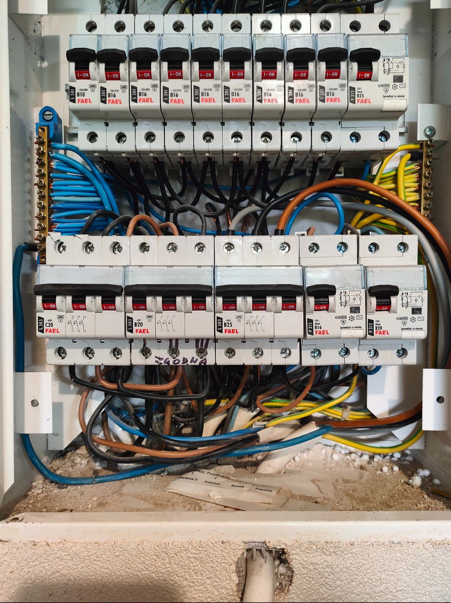 Rozdzielnica elektryczna z bezpiecznikami FAEL, widoczne przewody w różnych kolorach. Instalacja wewnątrz obudowy, z elementami montażowymi i oznaczeniami.