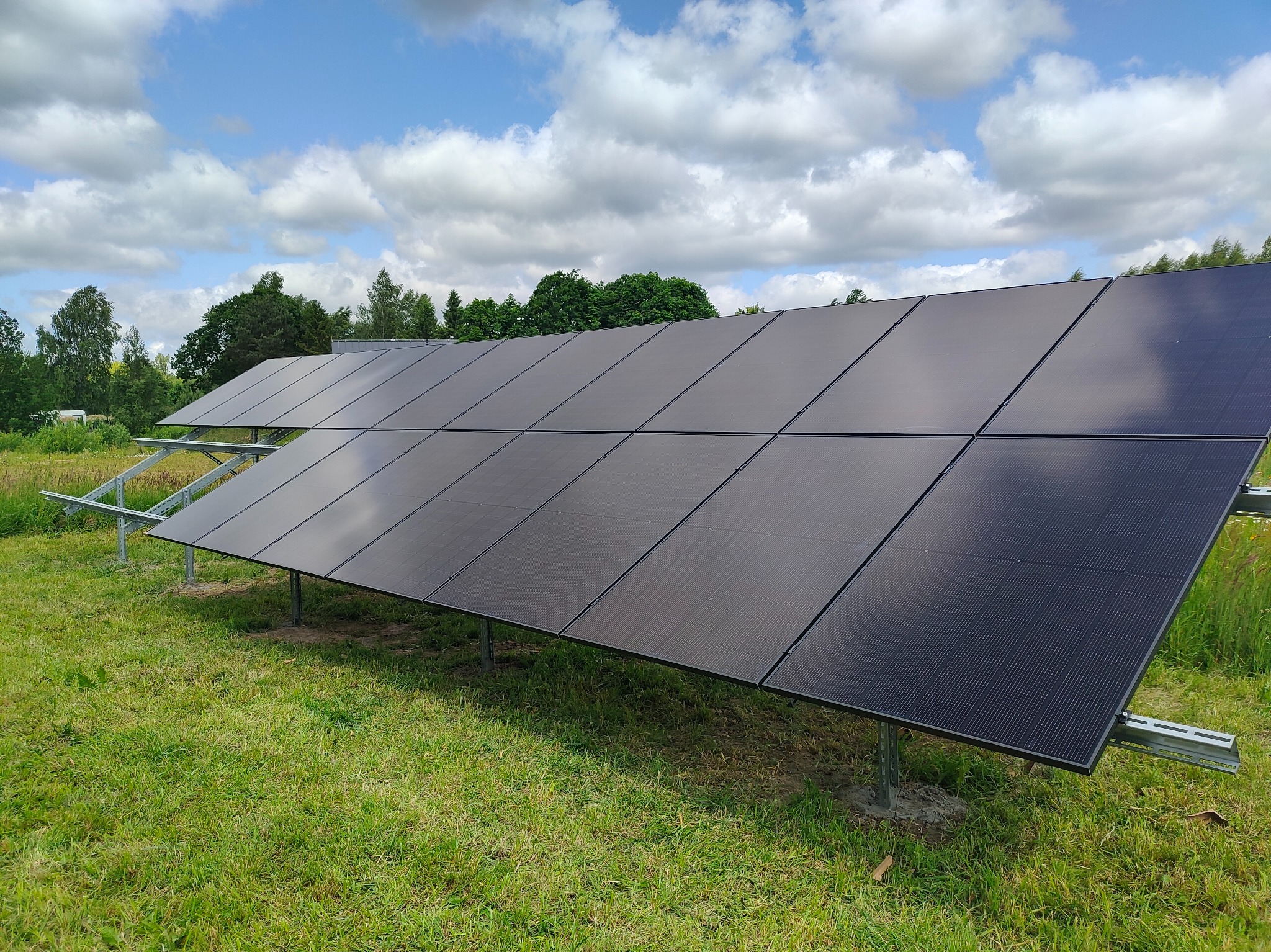 Rząd paneli fotowoltaicznych na metalowej konstrukcji, zainstalowanych na trawiastym terenie. W tle błękitne niebo z chmurami i zieleń drzew.