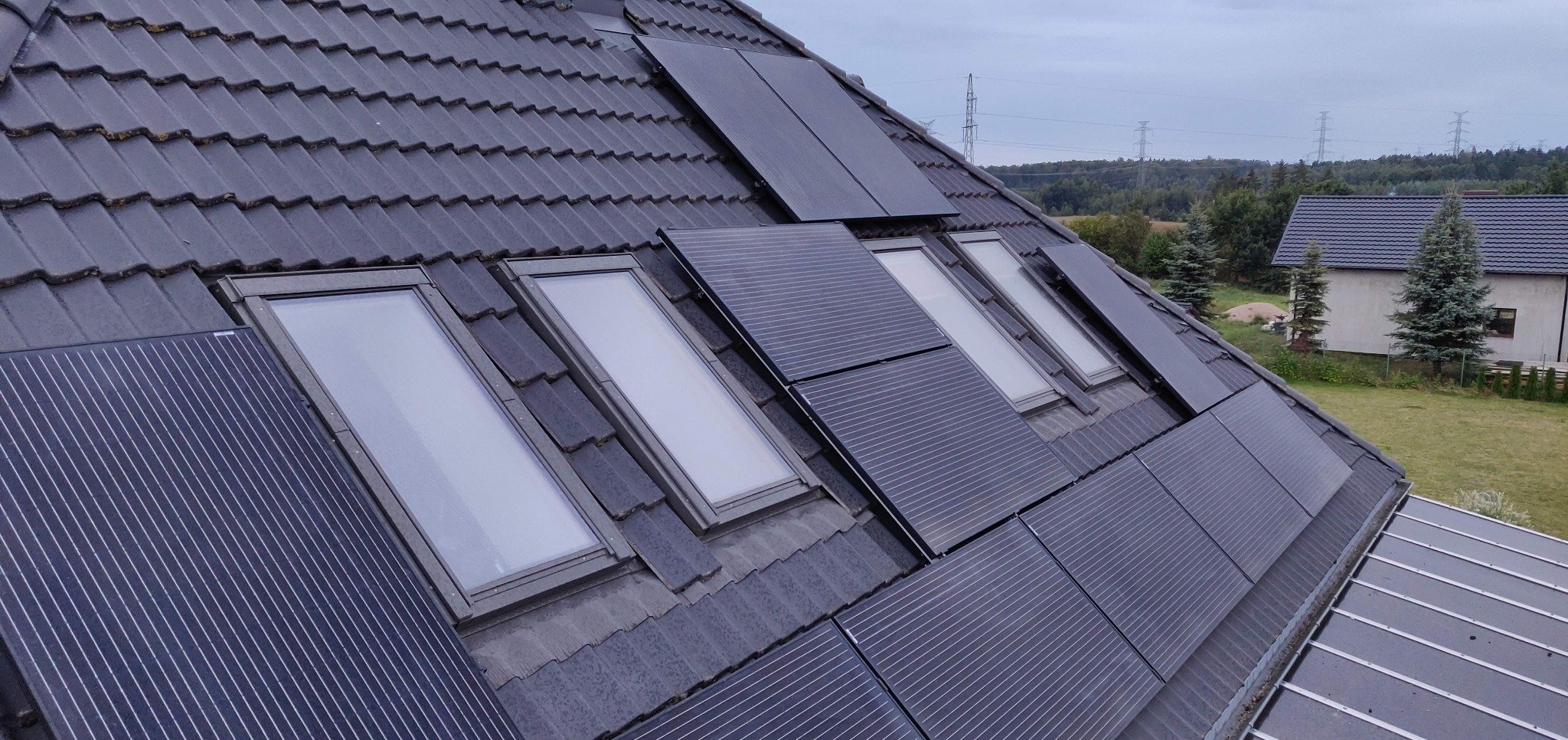 Czarne panele fotowoltaiczne zamontowane na dachu z dachówką ceramiczną, pomiędzy oknami dachowymi. W tle widoczne drzewa i słupy wysokiego napięcia.